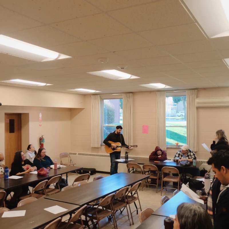 A group of people in a community hall listen as a man stands and plays an acoustic guitar for them during a gathering.