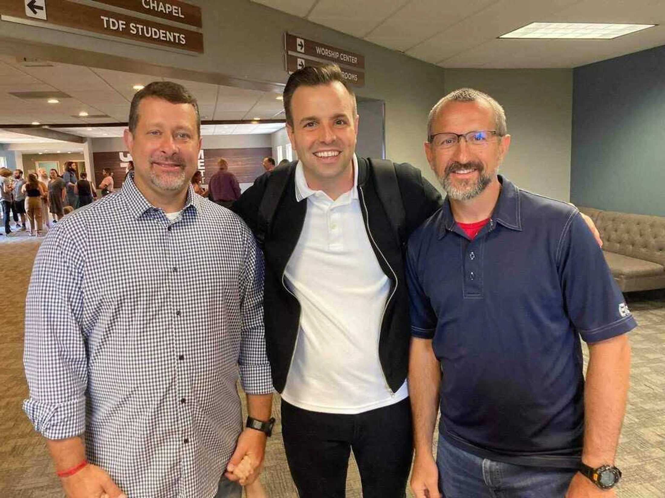 Three men stand side-by-side, smiling for a photo in a brightly lit hallway. The man in the center has his arms around the other two.