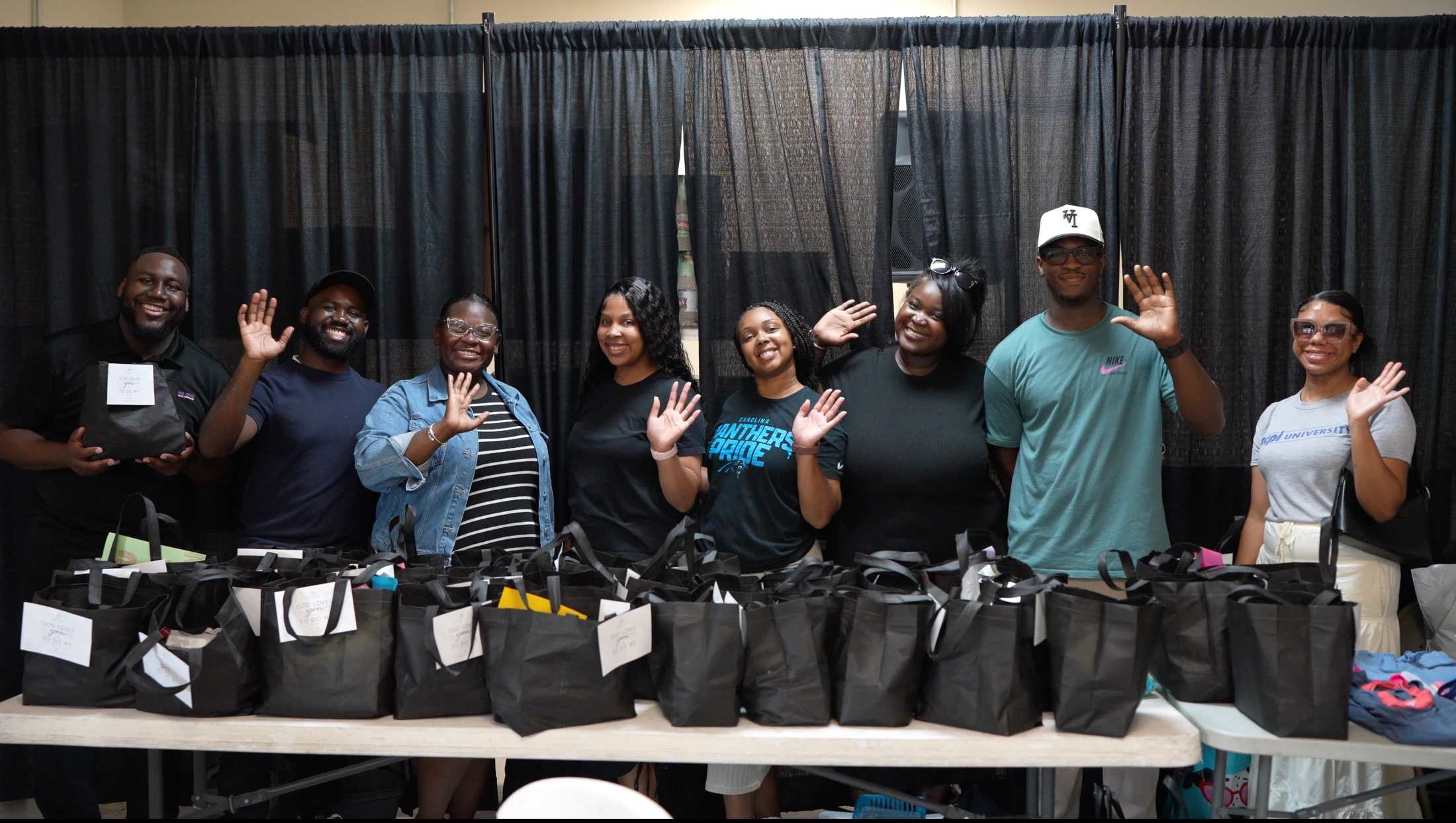 A group of eight smiling Black adults stand behind a long table filled with black gift bags, waving at the camera in front of a black curtain.