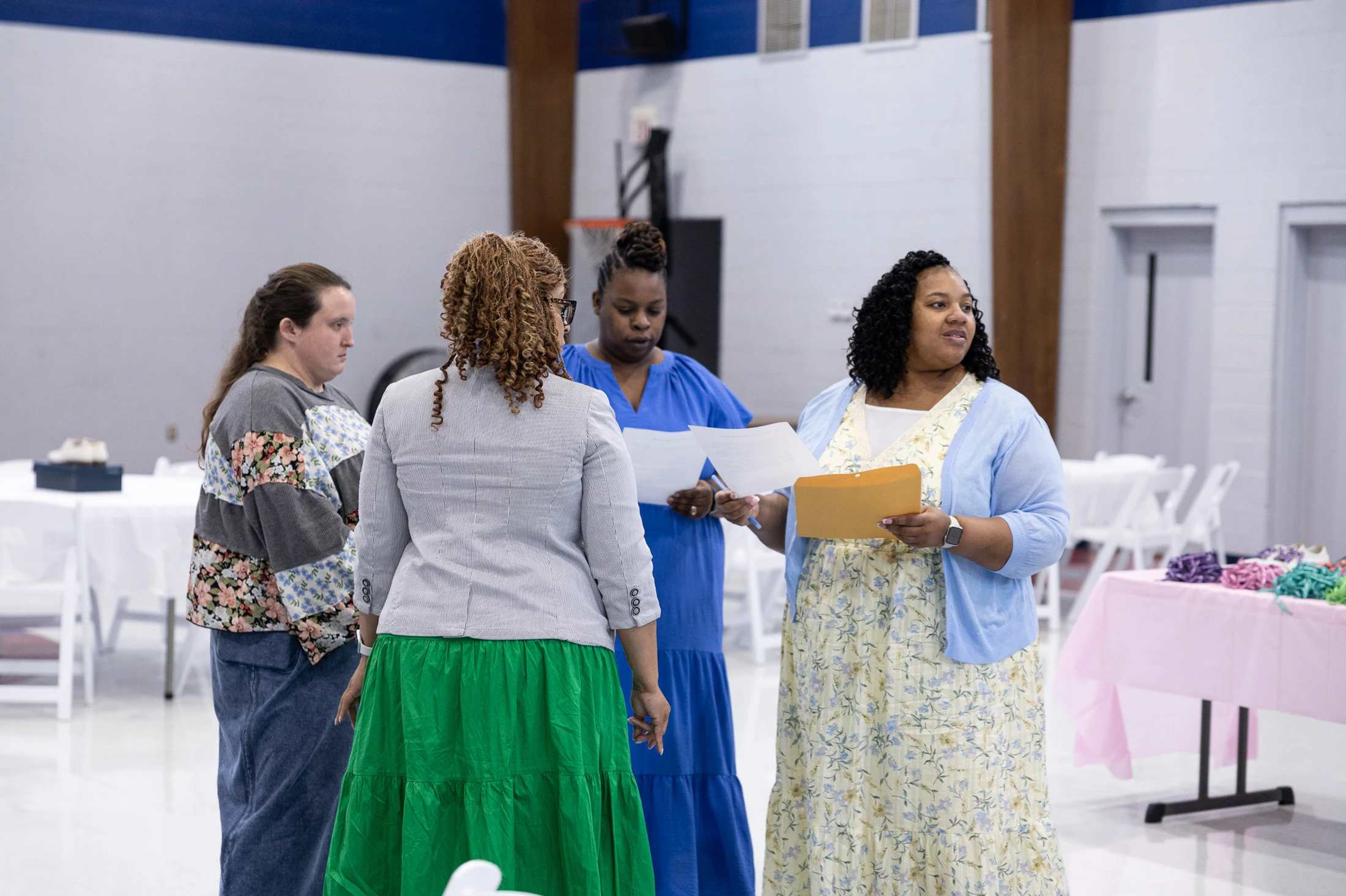 Four women stand together in a large hall with tables and chairs, appearing to be in a discussion. One woman holds a manila folder and papers.