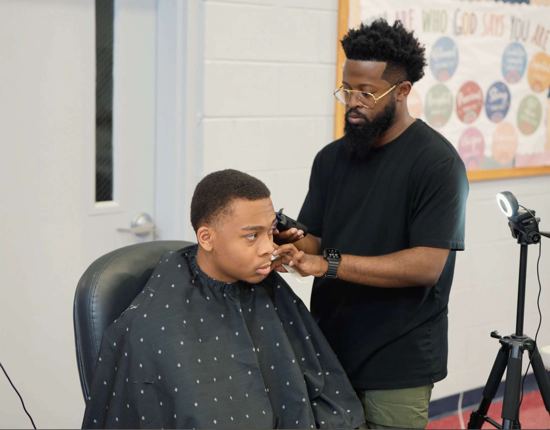 A barber with a beard and glasses carefully uses clippers to shape up a young man's hairline as he sits in a barber's chair wearing a black cape.
