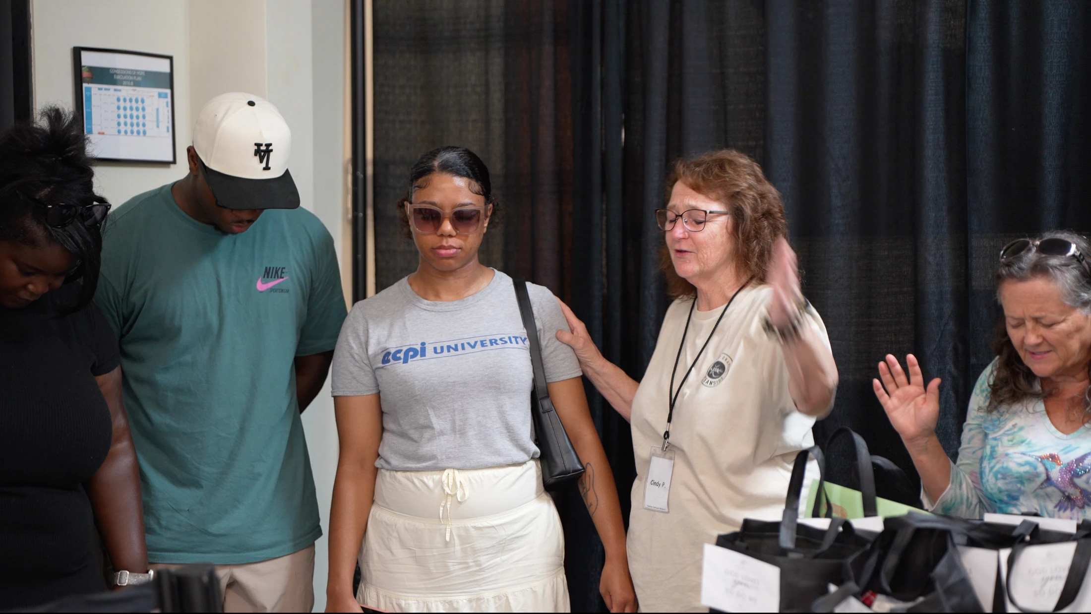 An older woman with glasses leads a diverse group of people in prayer, placing a hand on the shoulder of a young woman with her eyes closed.