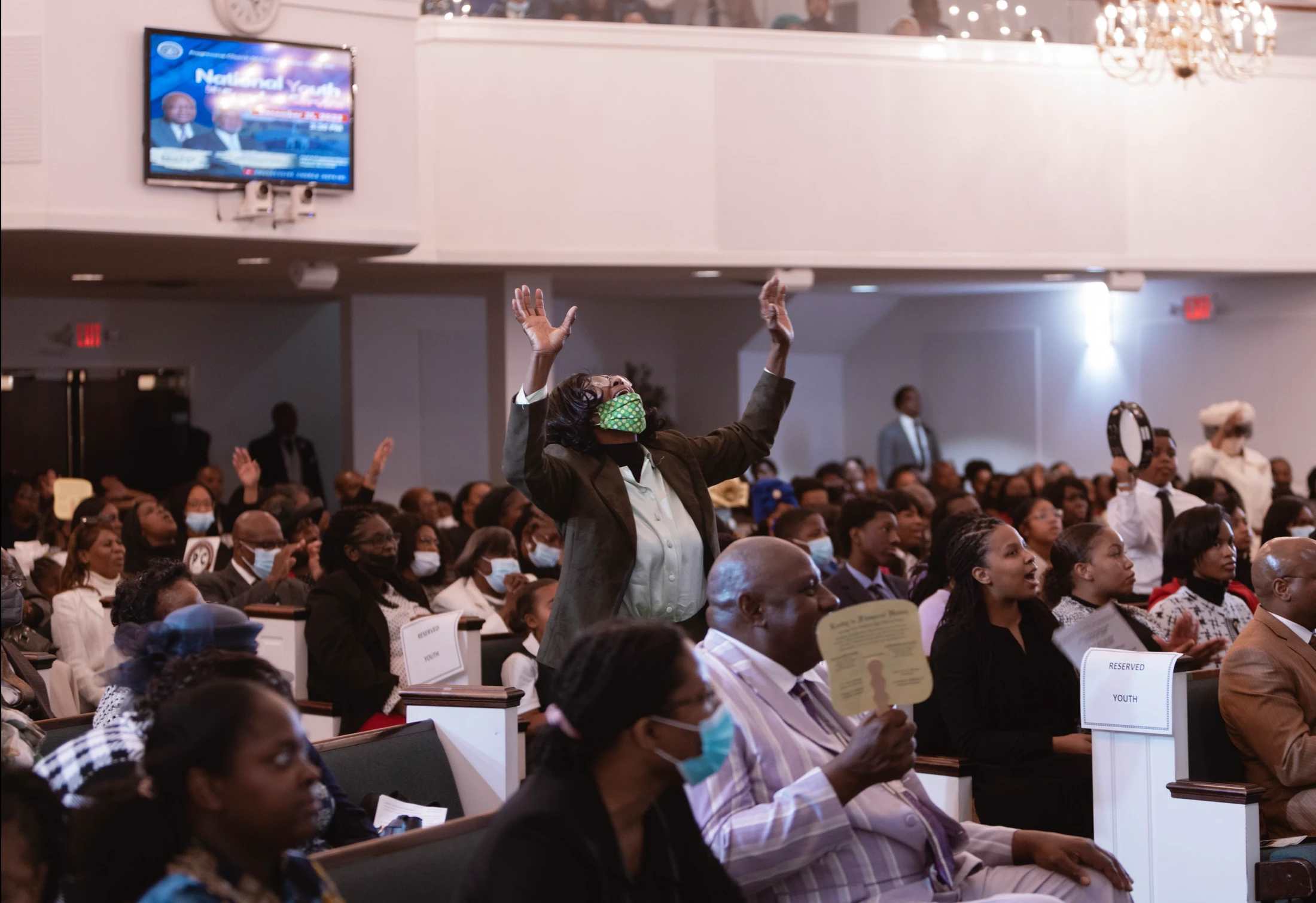 A Black woman stands in a church congregation with her hands raised in worship. Many other people are seated in the pews around her, some wearing masks.