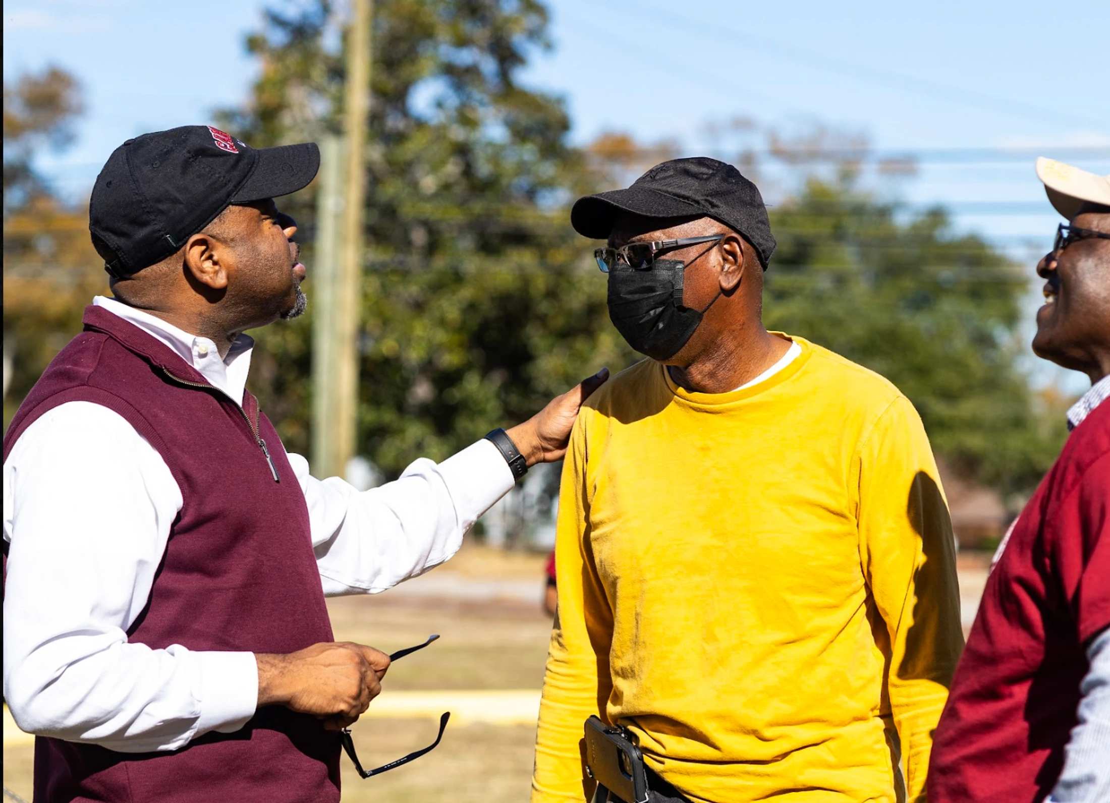 Three Black men talking outdoors. One man in a maroon vest has his hand on the shoulder of a man in a yellow shirt and face mask.