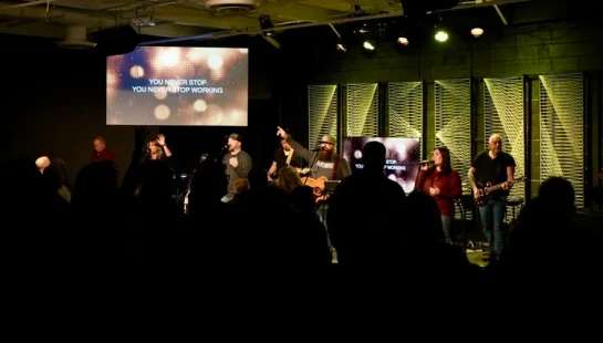 A worship band on a dimly lit stage with yellow lights, leading a silhouetted congregation. A screen shows the lyrics 'You Never Stop Working'.