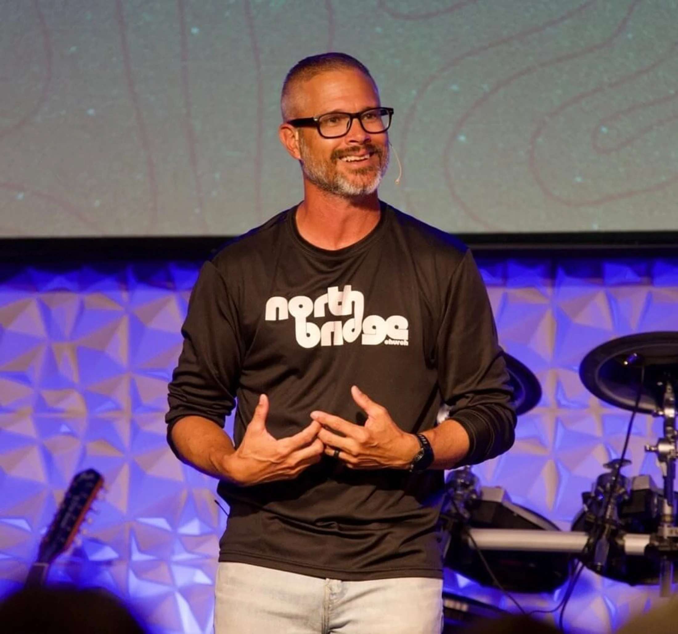 A smiling man with glasses and a beard, wearing a black North Bridge Church shirt, gestures with his hands while speaking on a stage with a blue background.