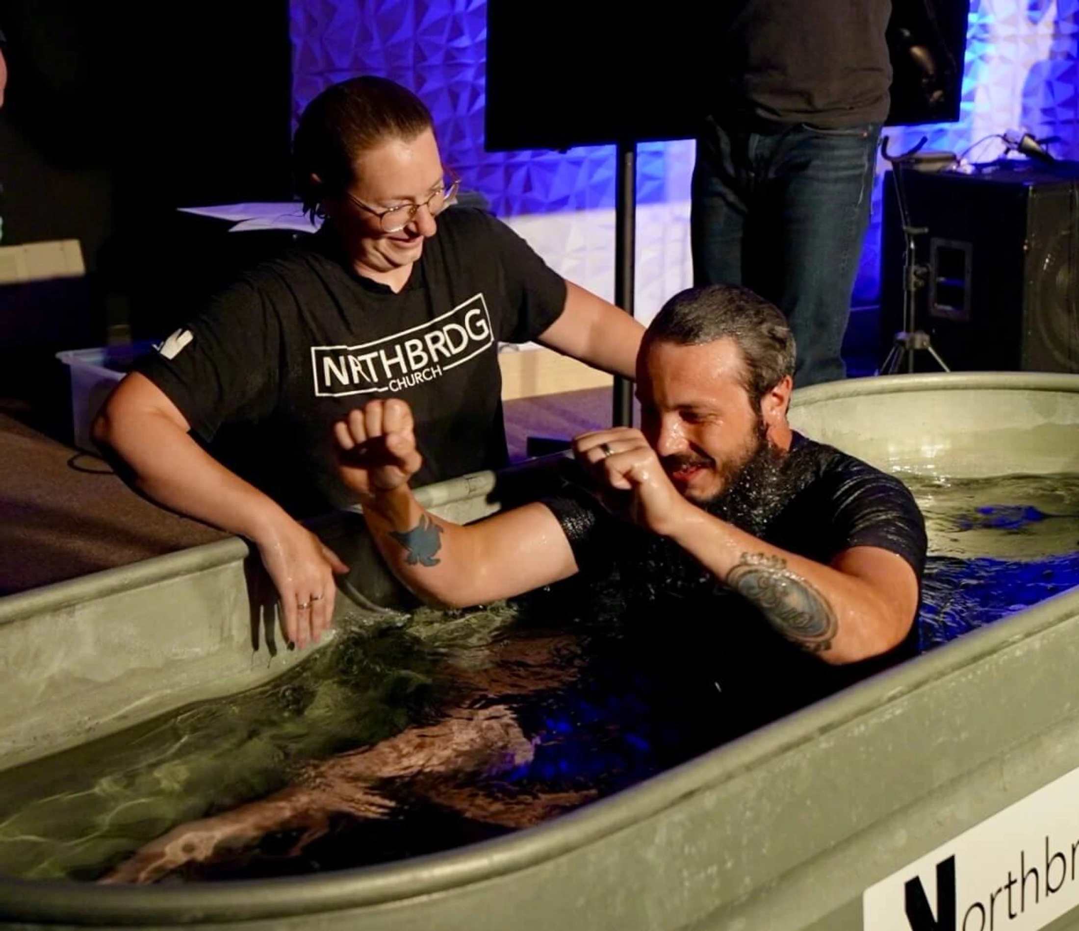 A smiling, bearded man with tattoos is helped out of a metal baptismal tub by a woman in a black t-shirt during a church service.
