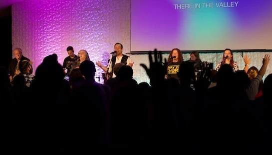 A worship band performs on a purple-lit stage in front of a silhouetted crowd with hands raised. A screen behind the band displays lyrics.