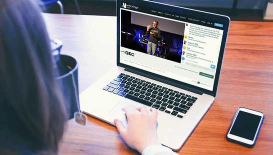 An over-the-shoulder view of a person watching a church service live stream on a laptop, with a video of a speaker and a live chat window visible on screen.