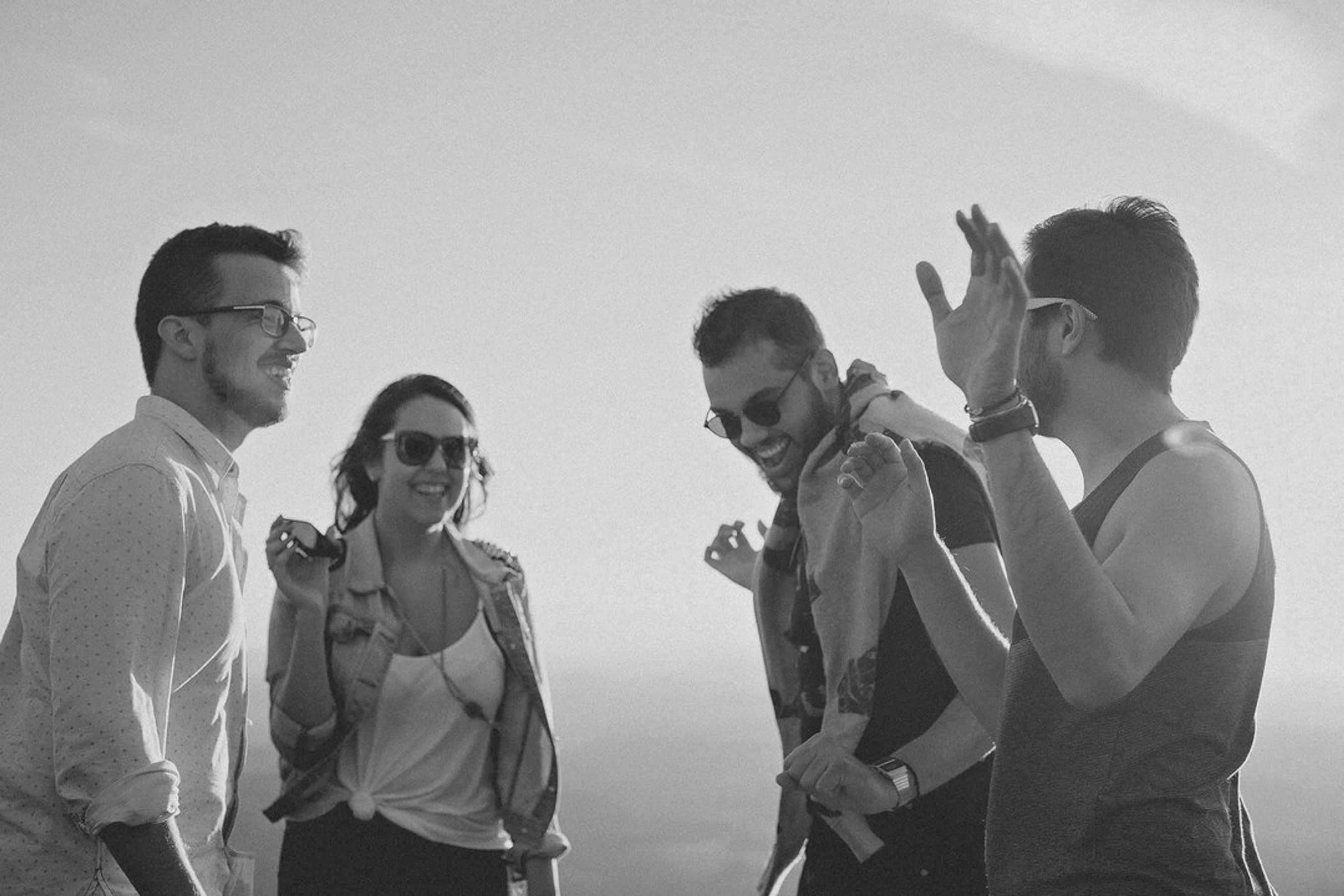 A black and white photo of a group of four friends laughing and talking together outdoors against a bright sky, conveying a sense of happiness and camaraderie.