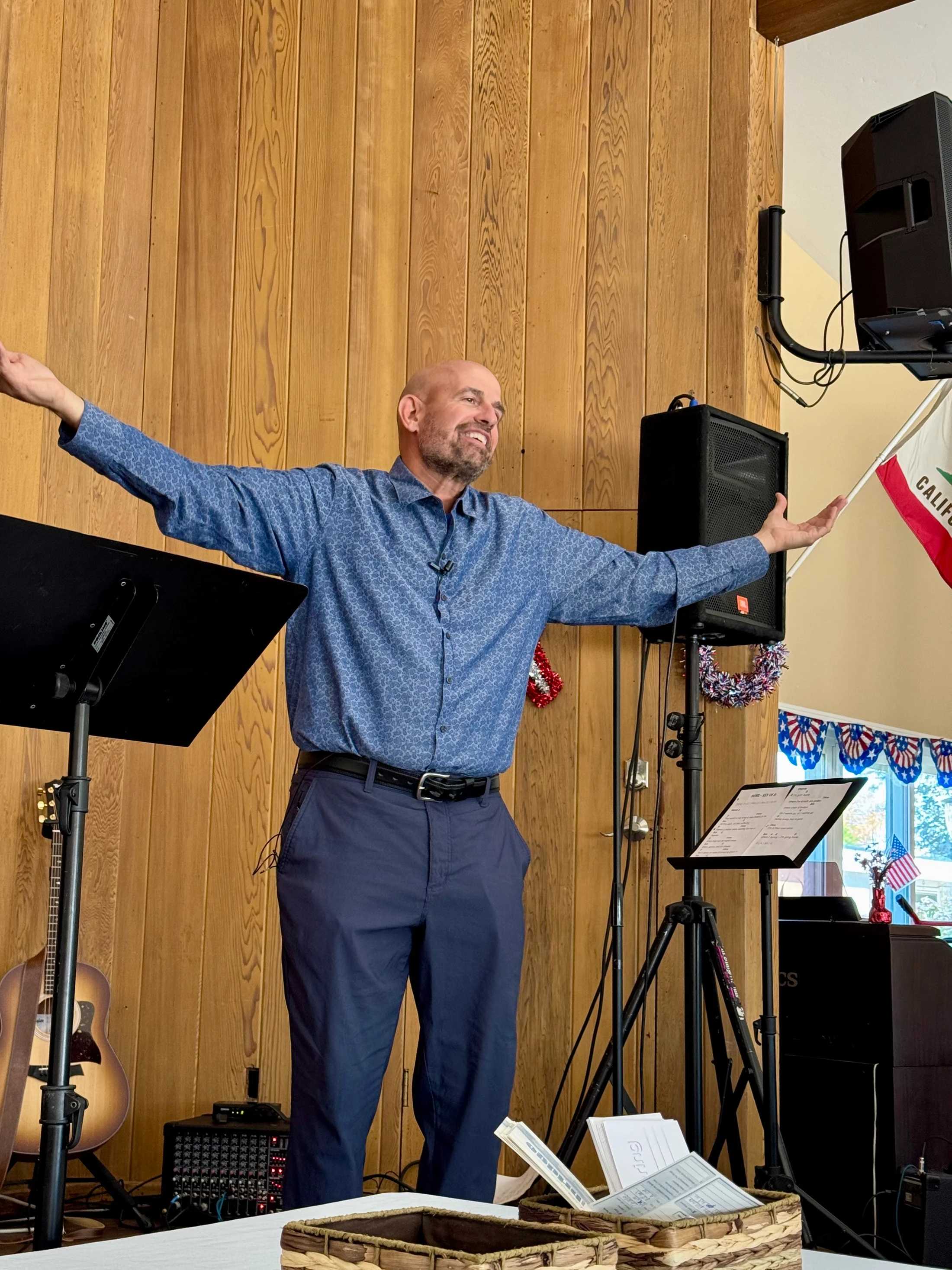 A bald man with a beard smiles as he speaks on a stage with a wood-paneled wall, his arms outstretched. He is surrounded by audio equipment and patriotic decor.