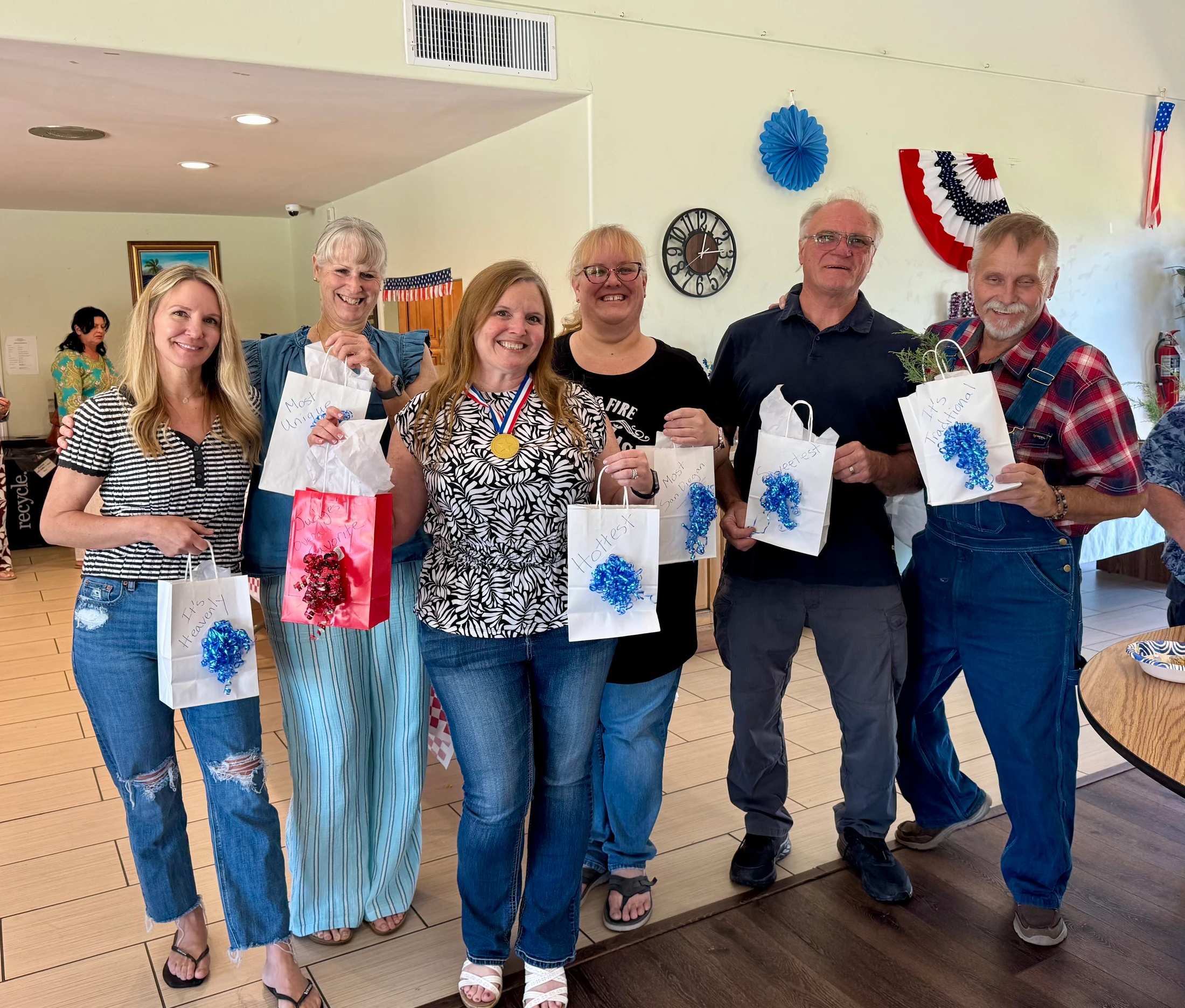 A group of six smiling adults pose for a photo indoors, holding small gift bags. The woman in the center proudly wears a gold medal around her neck.