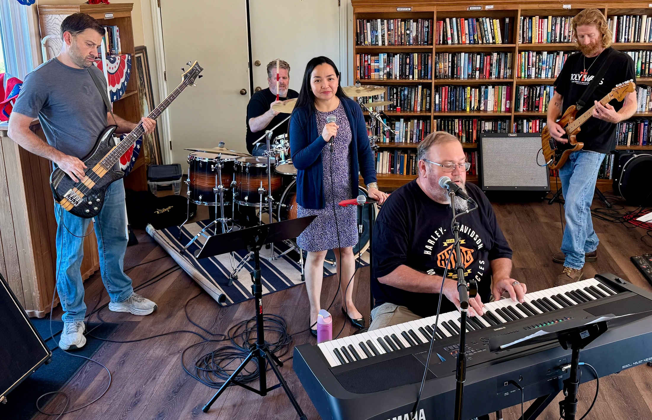 A five-person band rehearses in a room with a large bookshelf. A man plays keyboard and sings, a woman holds a mic, and men play bass, drums, and guitar.