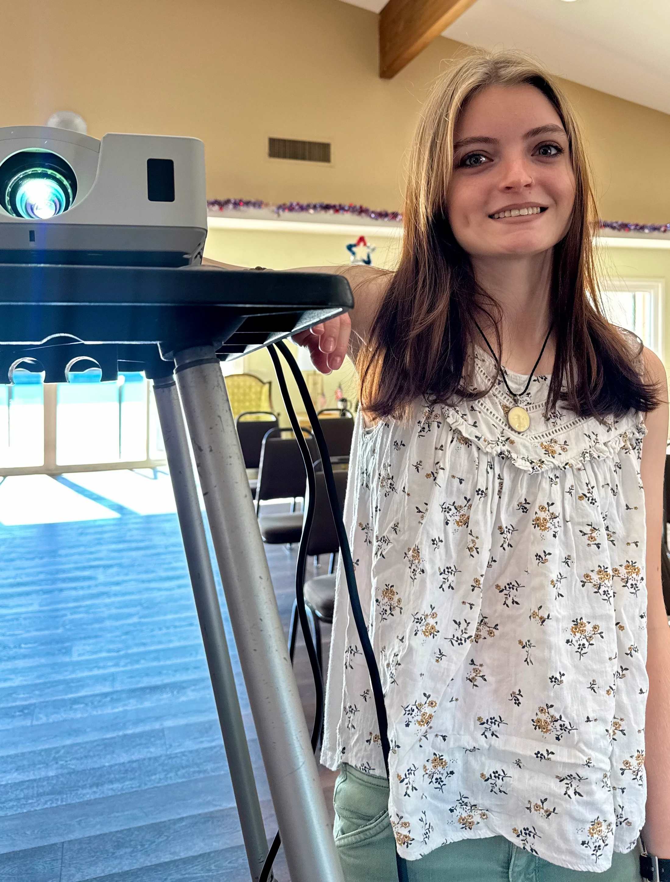 A young woman with long brown hair and a floral top smiles while standing in a room, with her hand resting on a lit video projector on a stand.