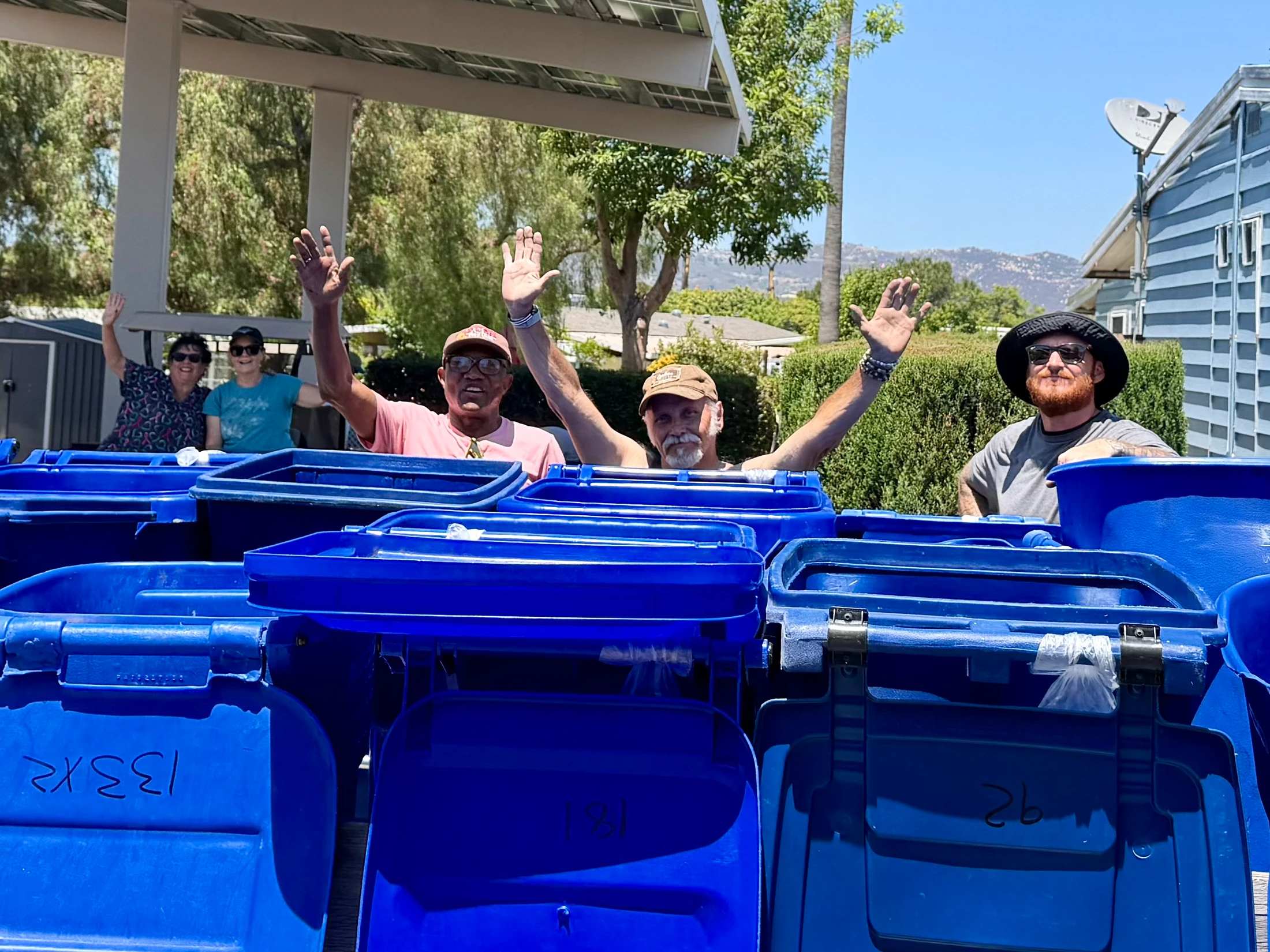 A group of five smiling people of various ages wave at the camera from behind a row of large, open, blue recycling bins on a sunny day outdoors.