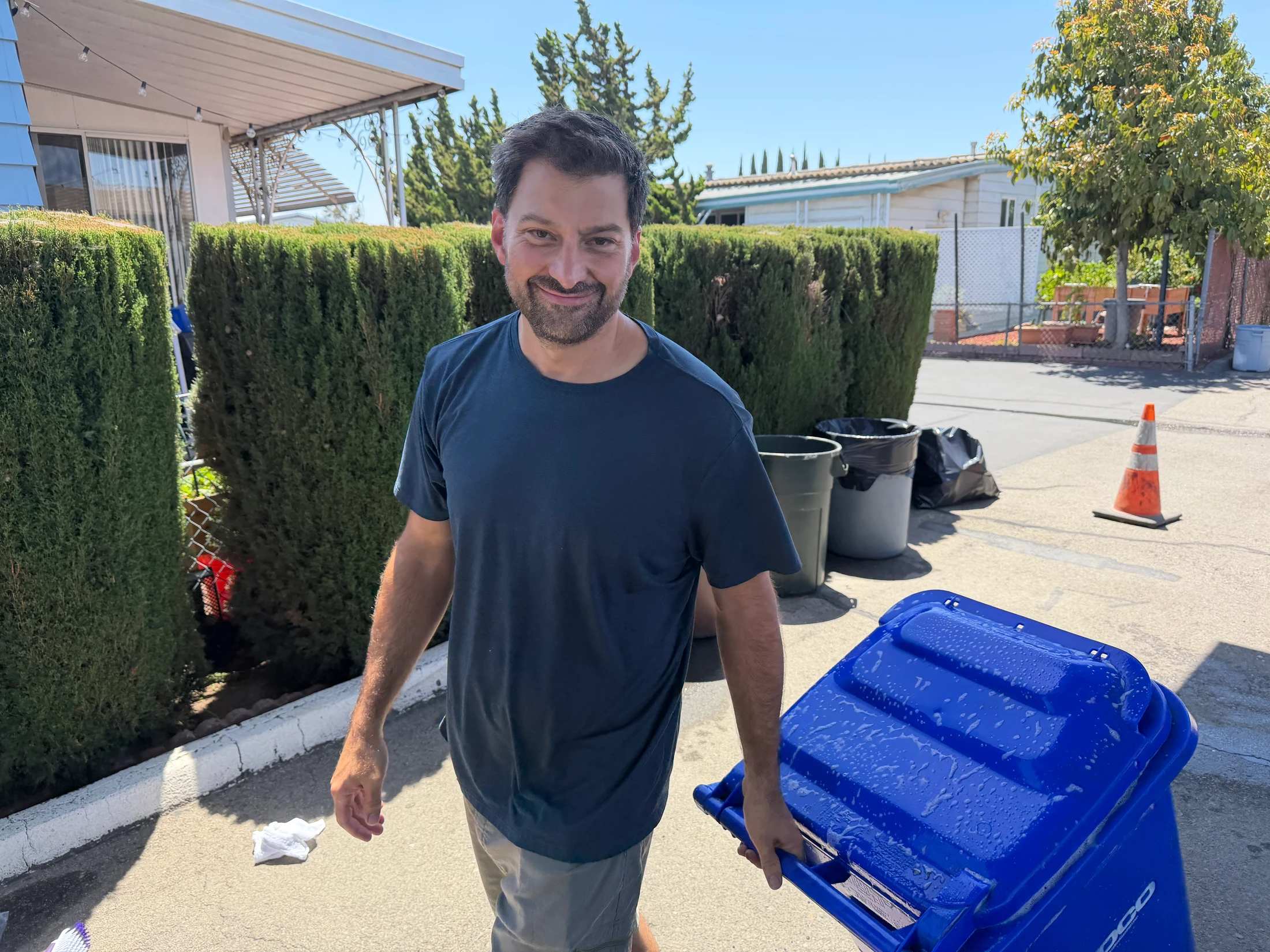 A man with dark hair and a beard, wearing a blue t-shirt, smiles as he stands outside on a sunny day holding the wet lid of a blue recycling bin.