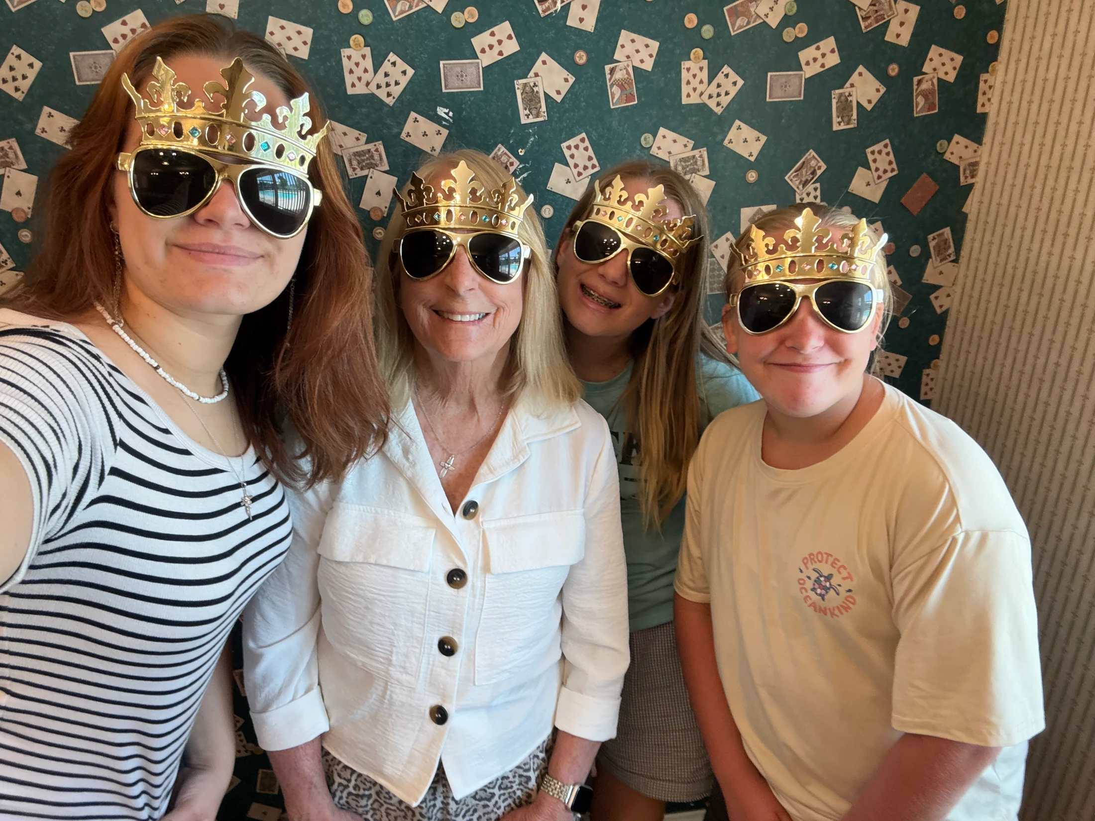 A selfie of a smiling woman and three children wearing matching golden crown sunglasses. They are posing in front of wallpaper with a playing card pattern.