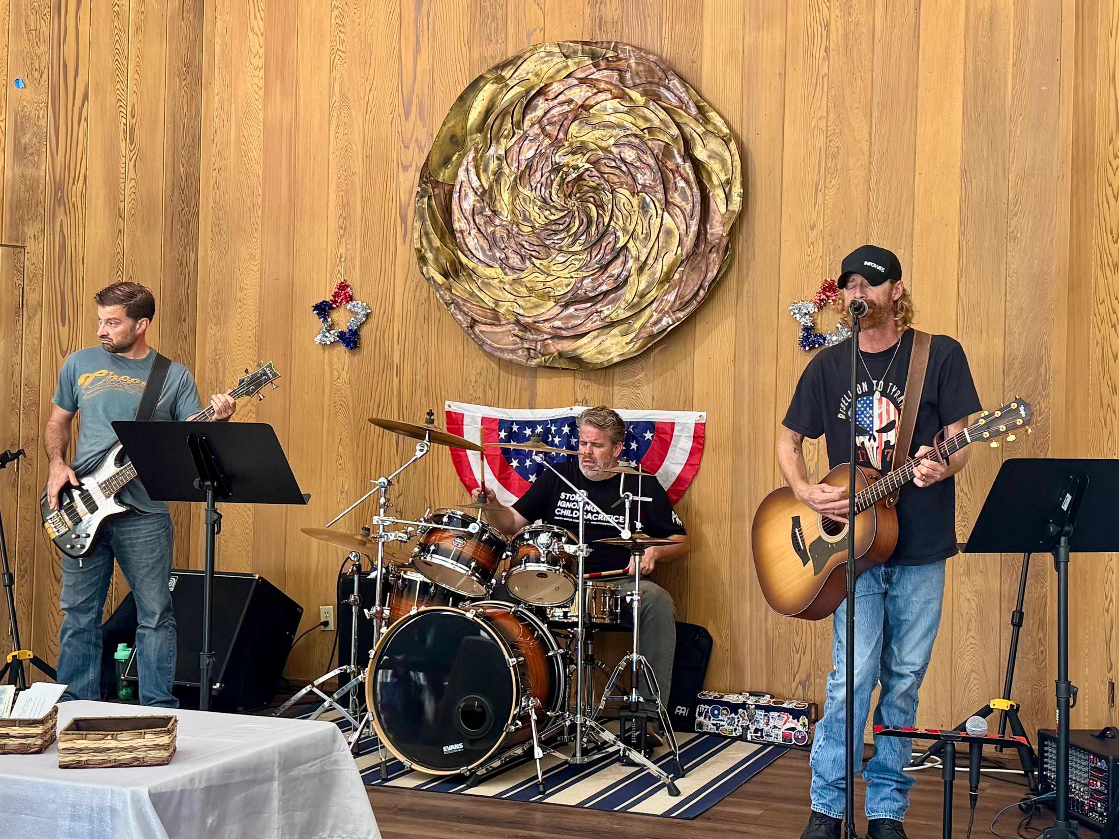 A three-person band plays on a small stage against a wood-paneled wall, featuring a guitarist/singer, a bassist, and a drummer.