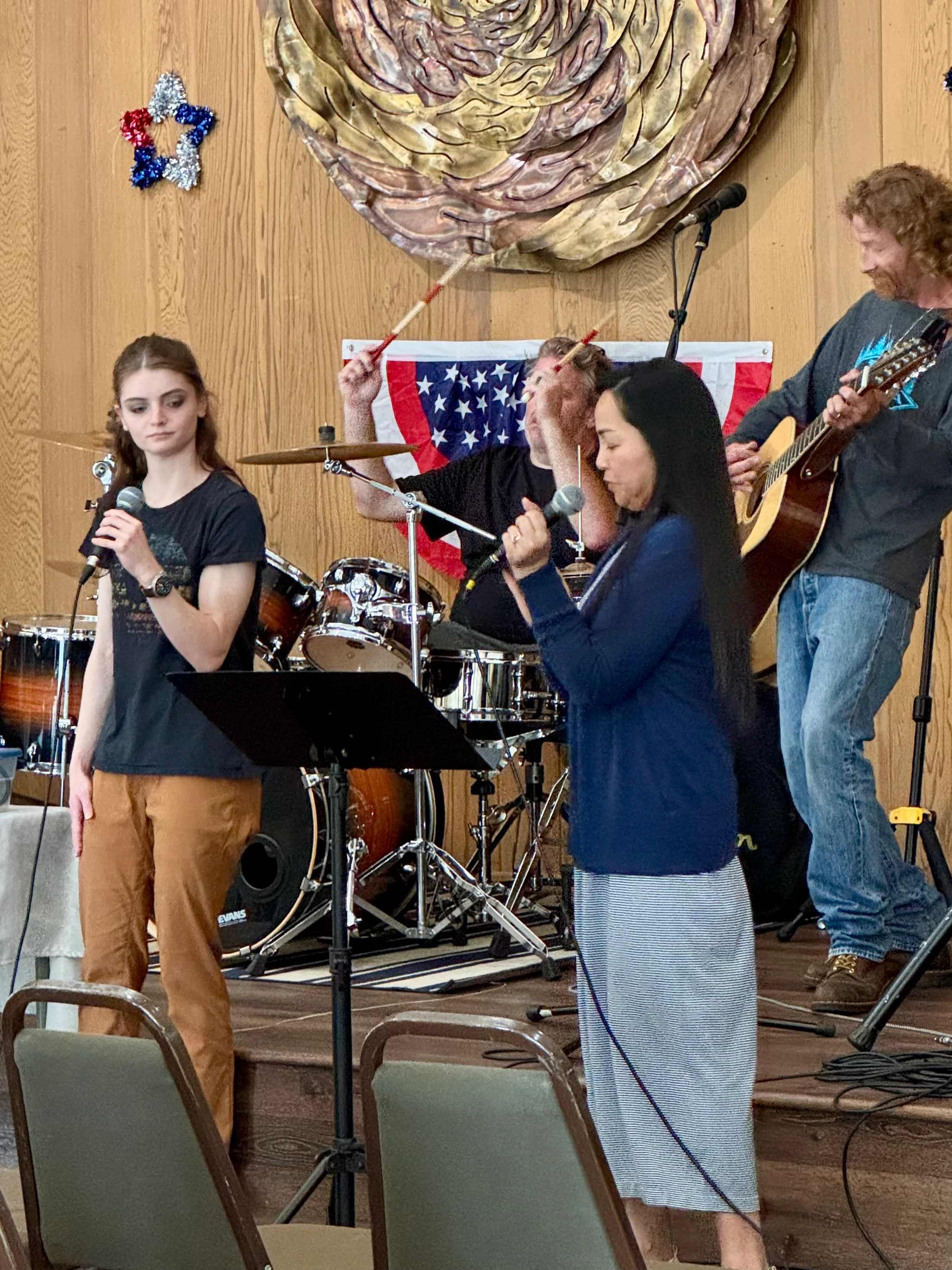A four-person band on a stage. Two women sing at microphones, a man plays guitar, and another plays drums. An American flag hangs on the wood-paneled wall.