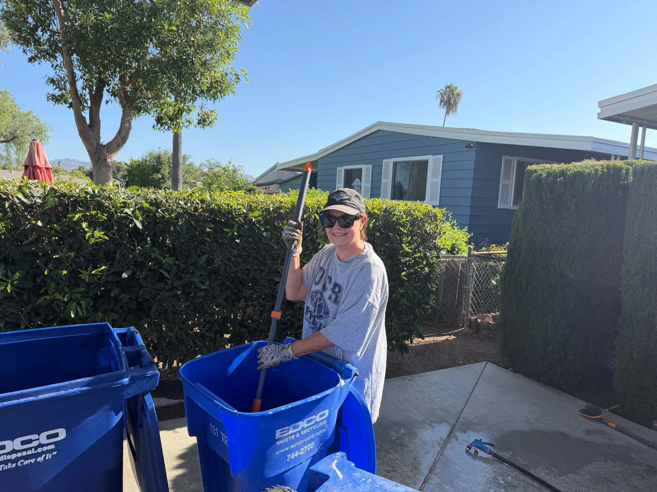 A smiling woman wearing a hat, sunglasses, and gloves uses a long-handled brush to clean the inside of a large blue recycling bin in a driveway.