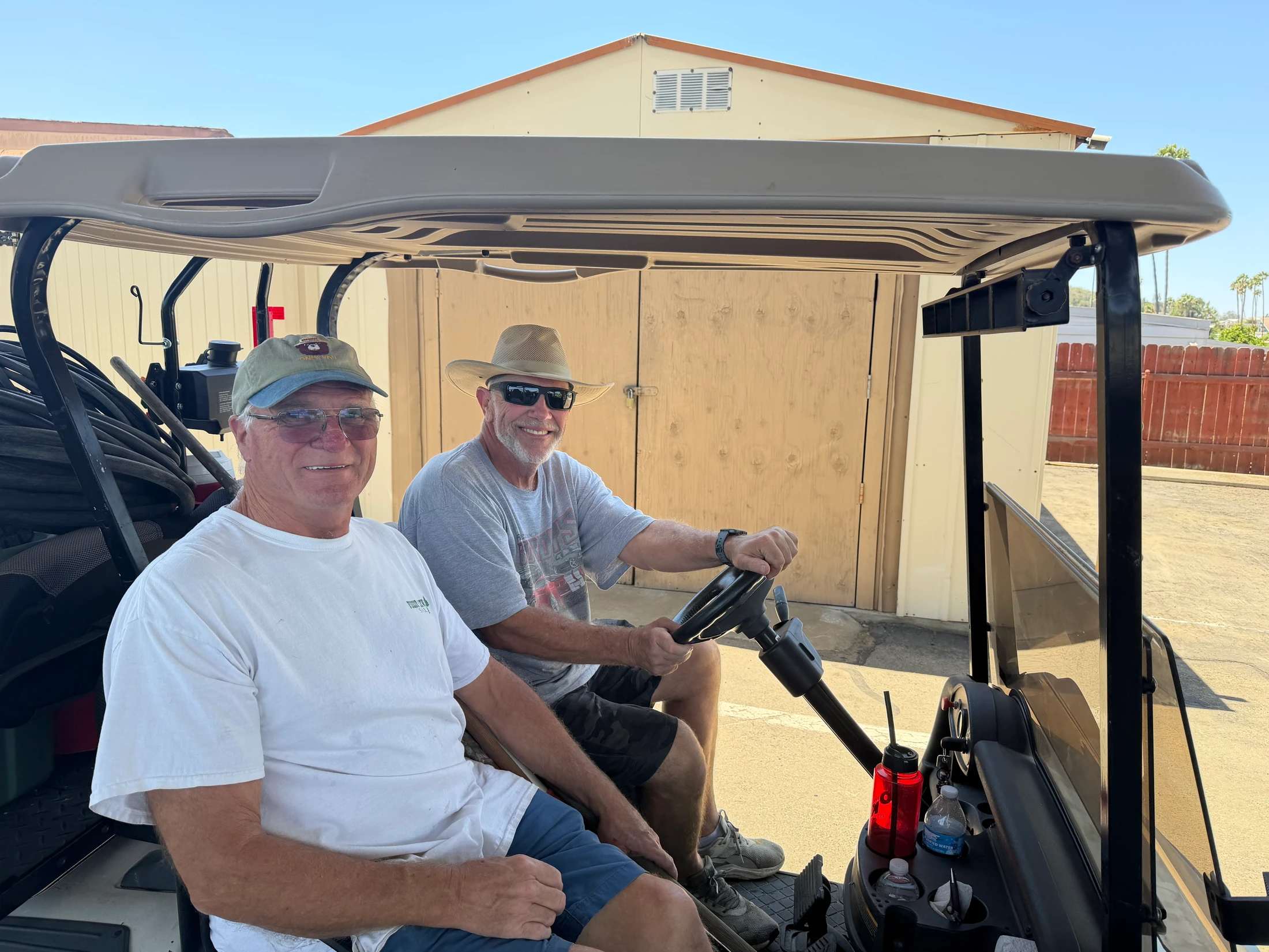 Two smiling older men sit in a golf cart on a sunny day. The man on the right is driving, wearing a hat and sunglasses, while the other man is a passenger.