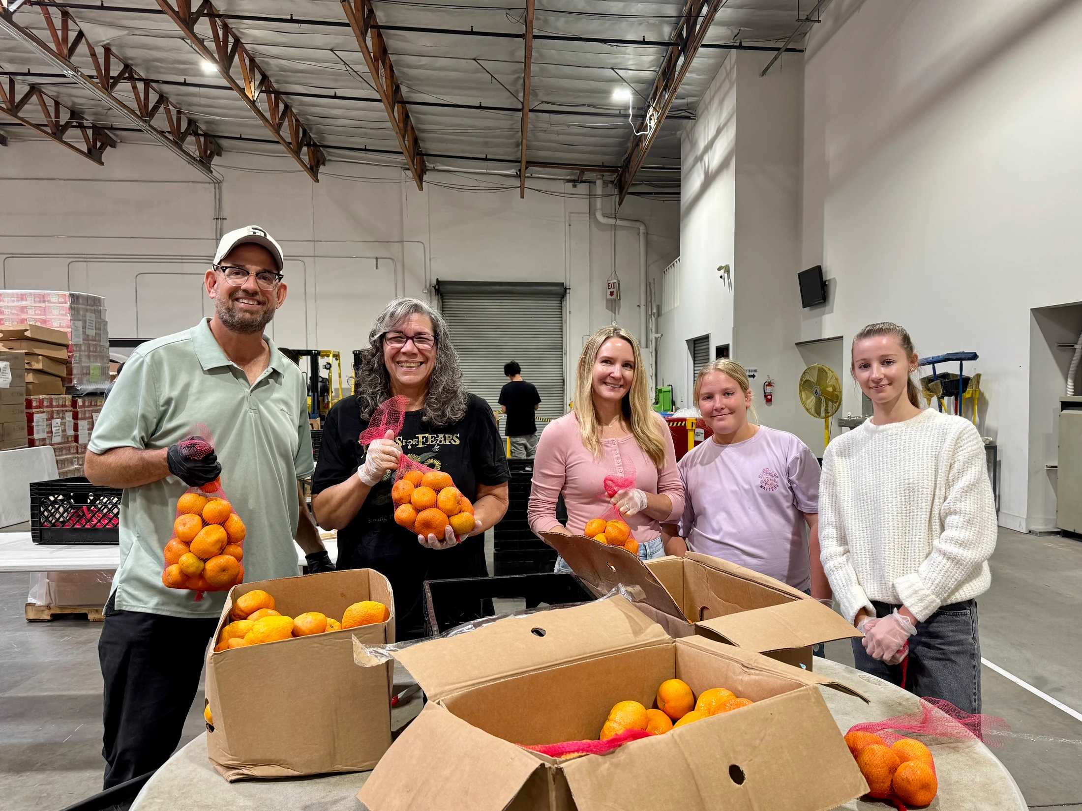 A group of five smiling volunteers stand in a warehouse, packing fresh oranges from cardboard boxes into red mesh bags for donation.
