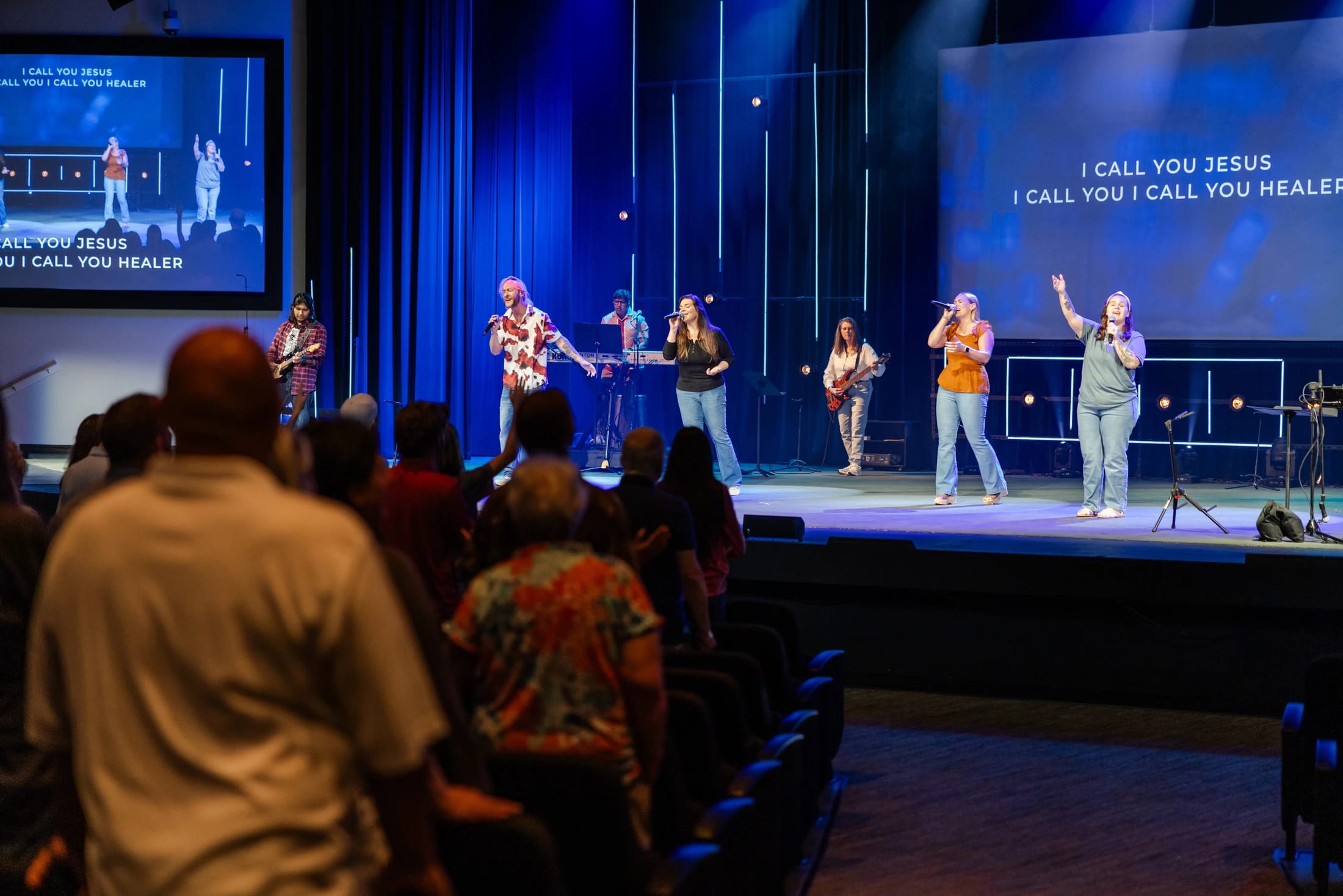 A view from the audience of a band performing on a stage with blue lighting. Screens behind them display the lyrics, 'I call you Jesus.'