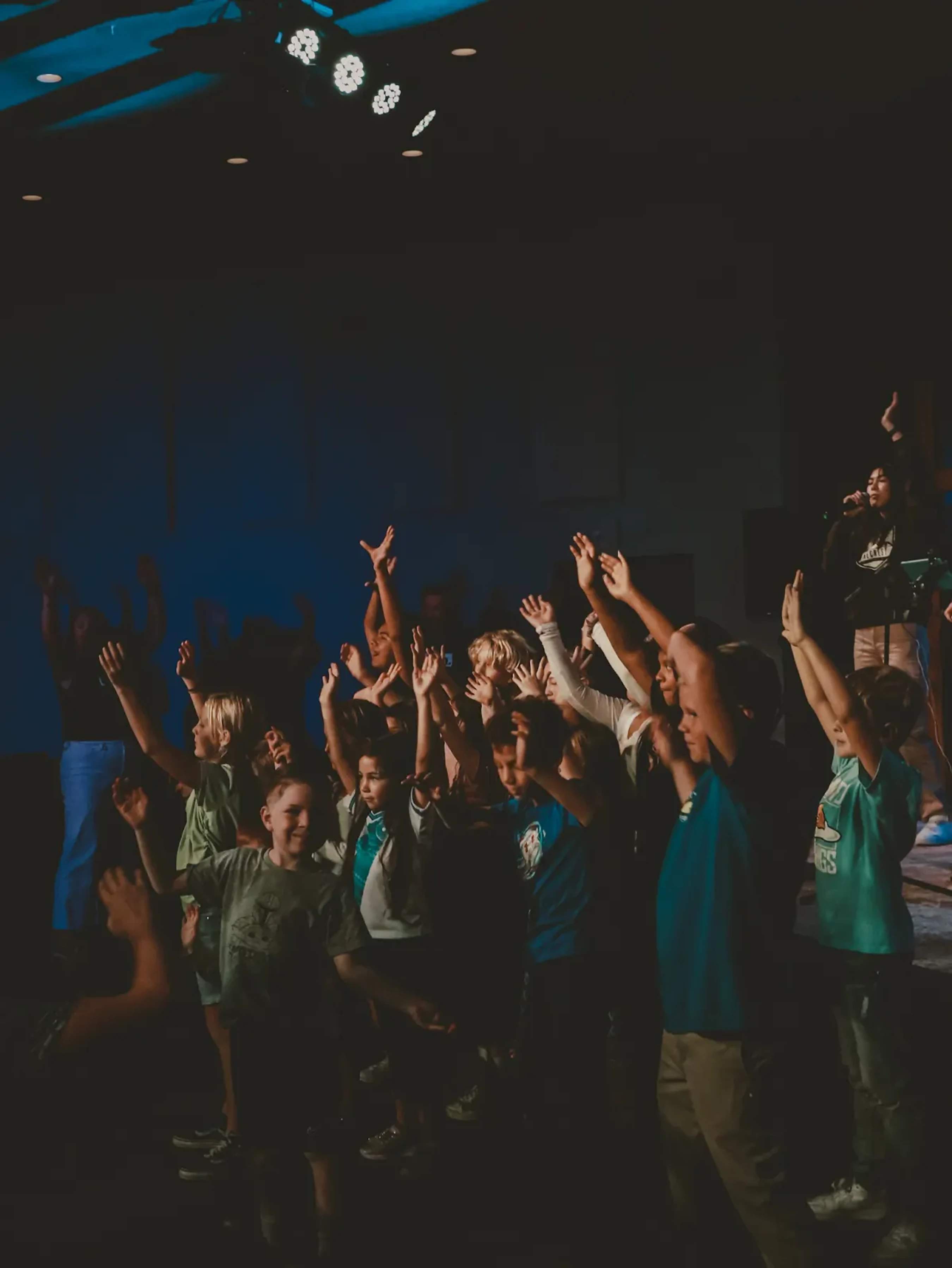 A group of diverse children smiling and raising their hands in a dimly lit room, with blue stage lights shining from above during a concert or service.
