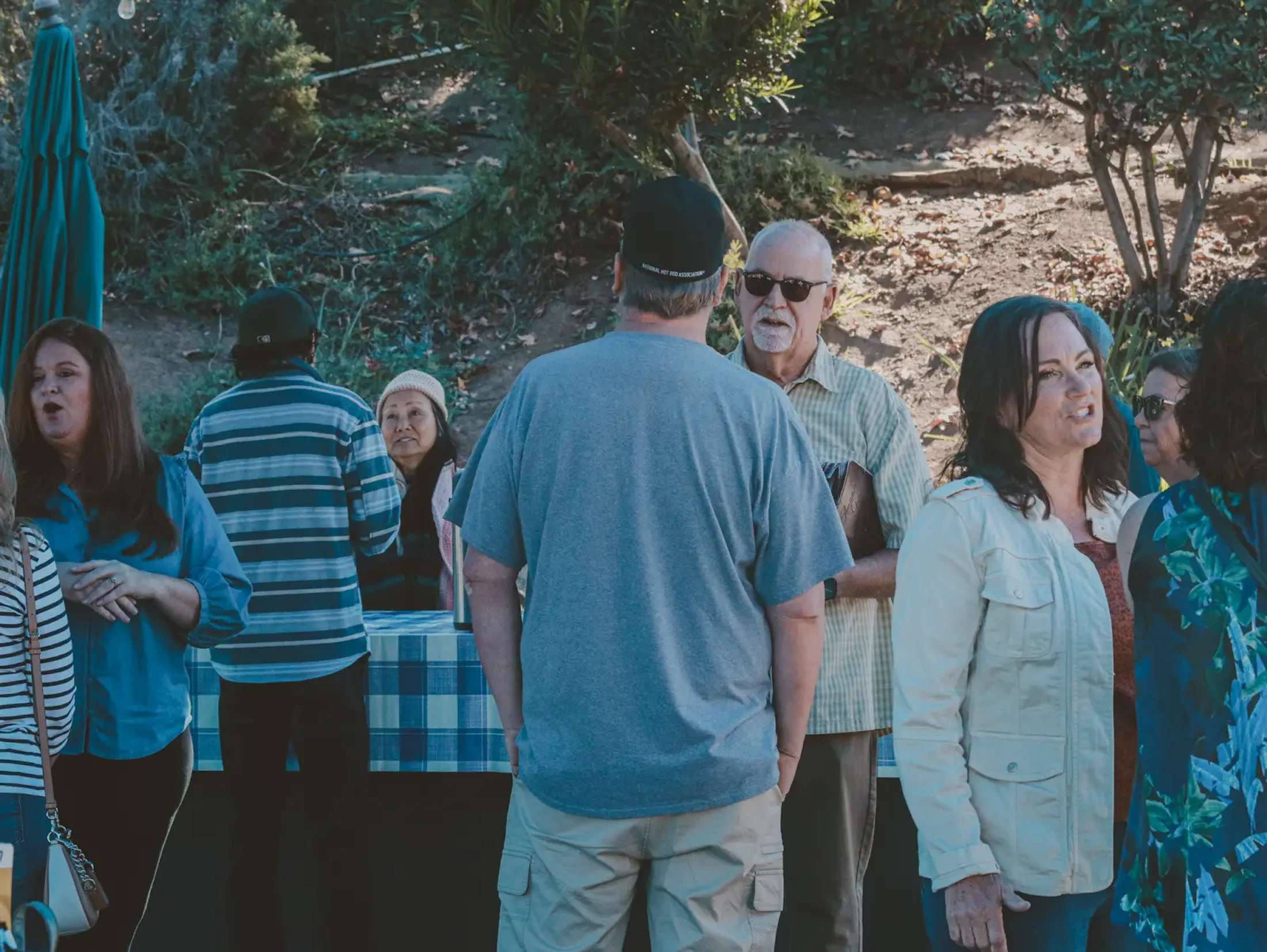 A diverse group of people standing outdoors in a casual setting, talking and mingling with each other near some trees and a table with a checkered cloth.
