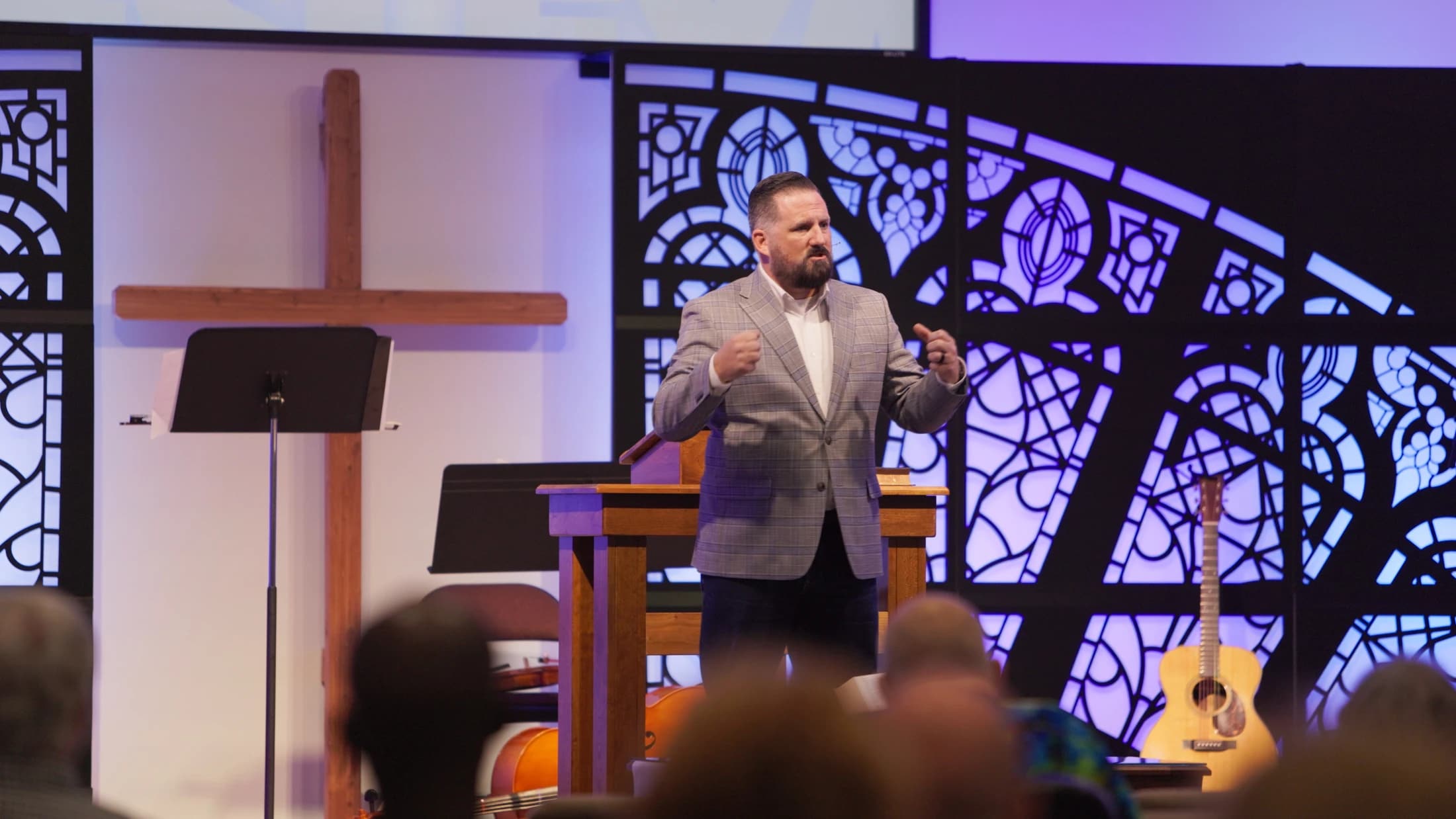 A man in a gray plaid blazer speaks from behind a wooden pulpit on a church stage. Behind him is a wooden cross and a modern, purple-lit stained glass design.