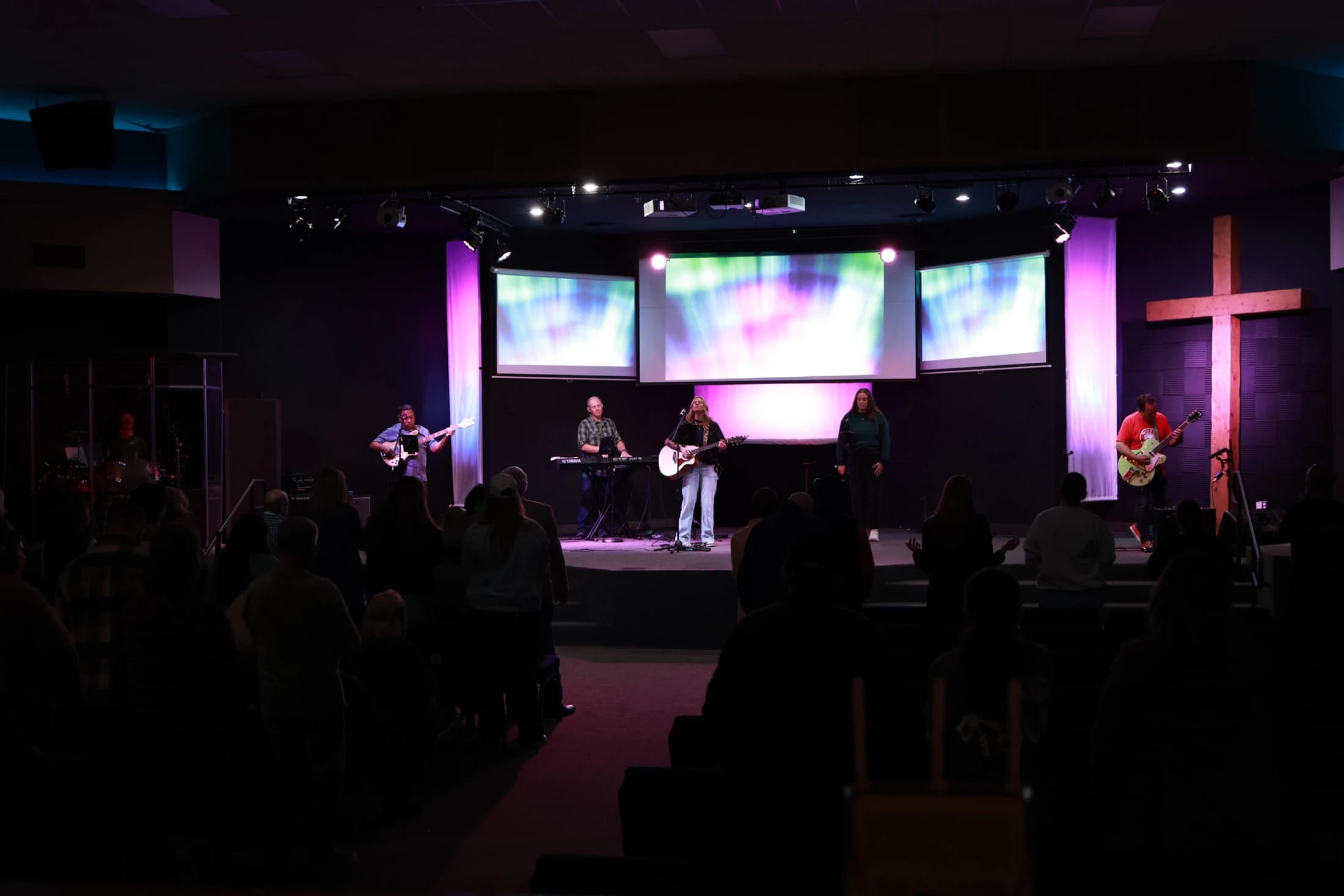 A band with singers and guitarists performs on a purple-lit stage in a church. A large wooden cross is on the right, and a silhouetted audience watches.
