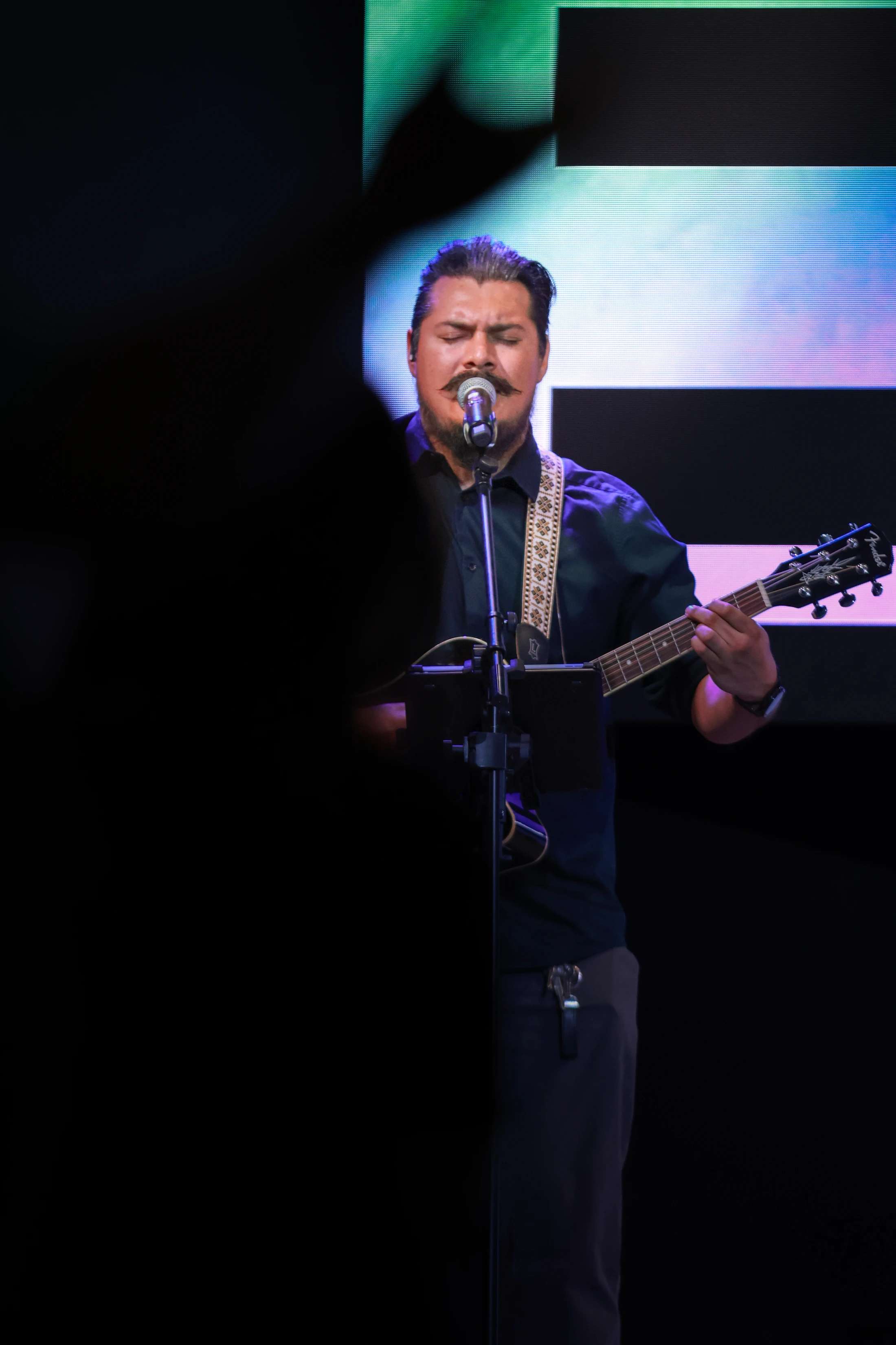 A man with a mustache sings passionately into a microphone while playing an acoustic guitar on a stage with a colorful screen behind him.