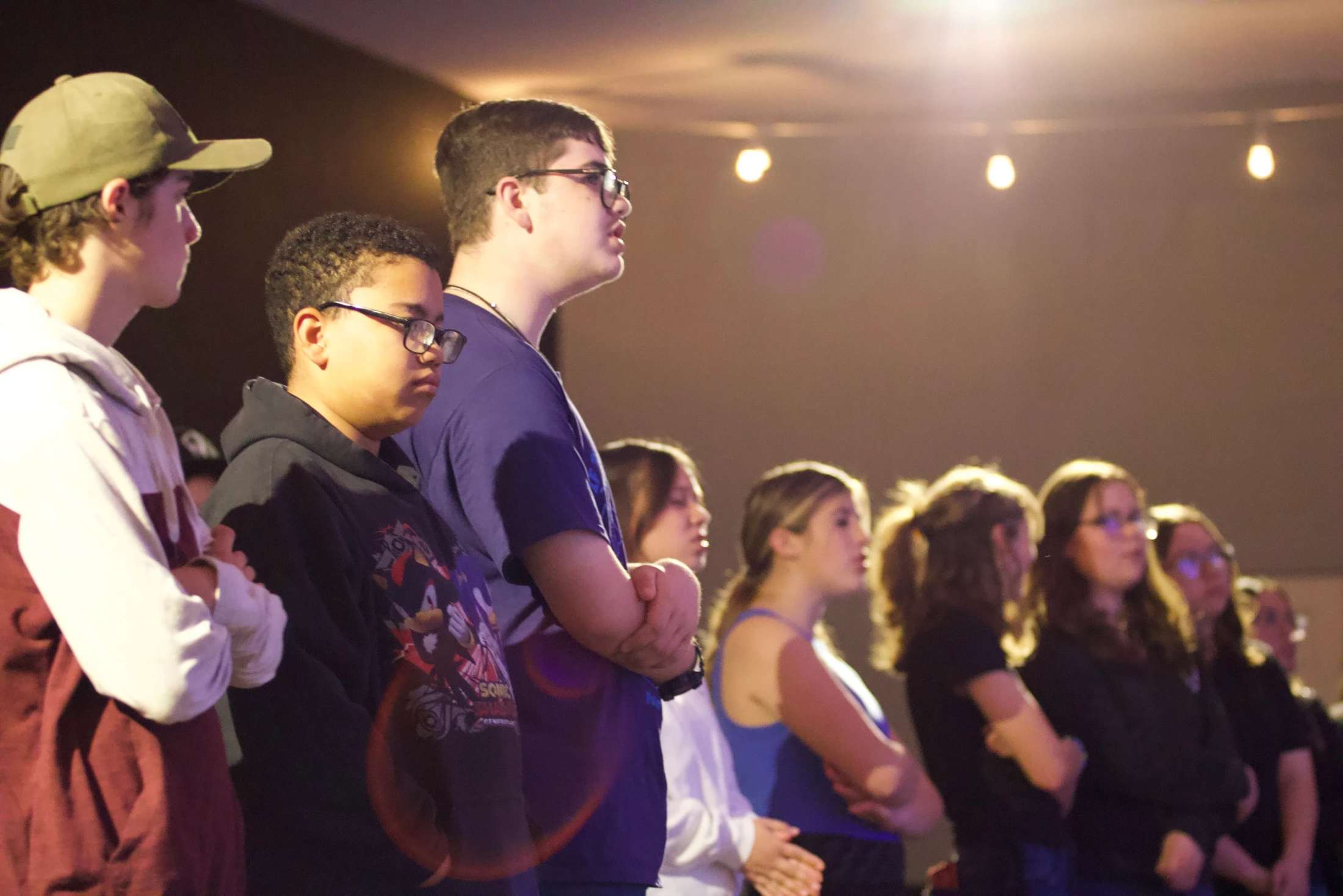 A diverse group of teenagers stands in a line in a warmly lit room, looking forward with focused expressions as they listen to something off-camera.