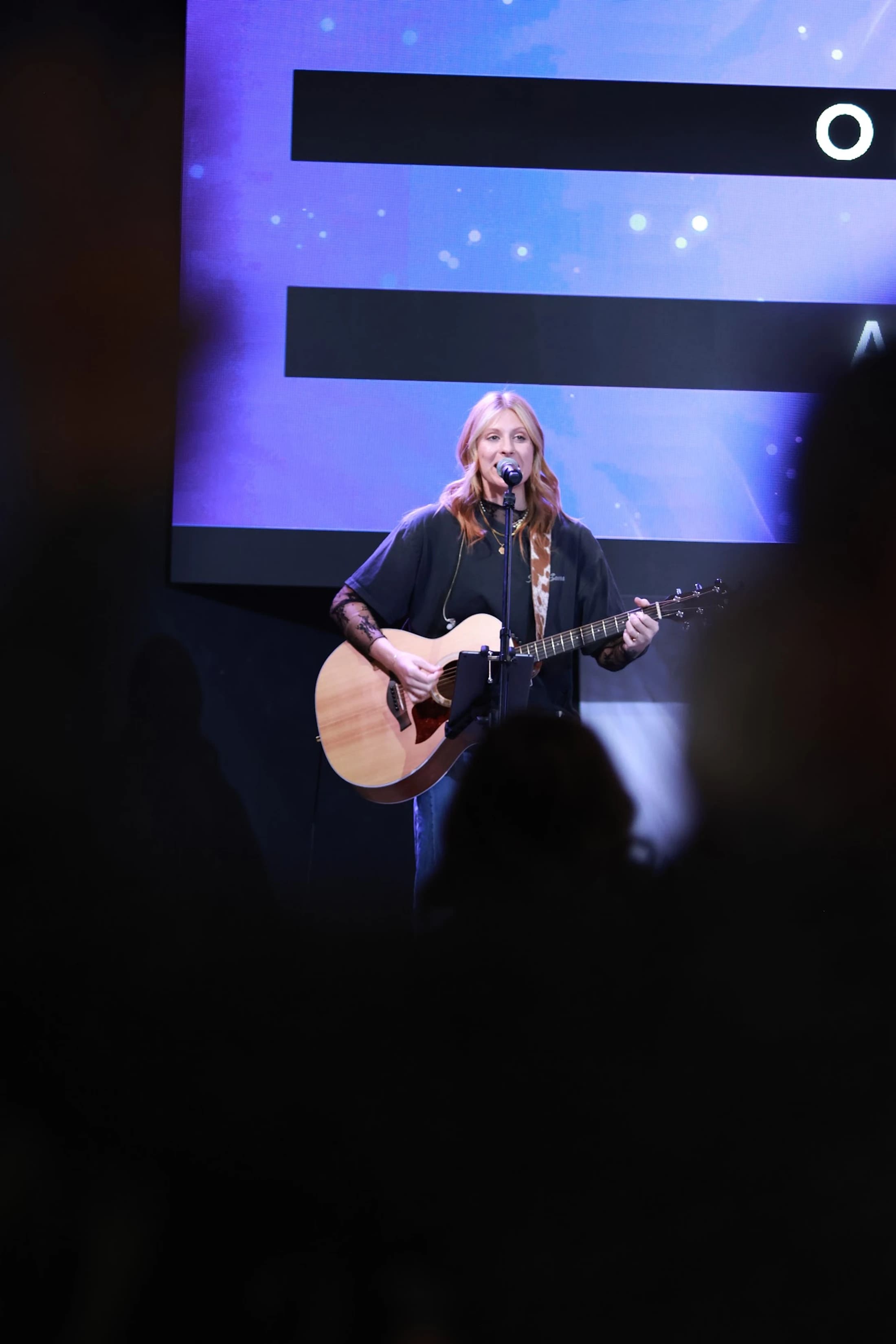 A woman with reddish-blonde hair sings into a microphone while playing an acoustic guitar on a dimly lit stage in front of a large purple screen.