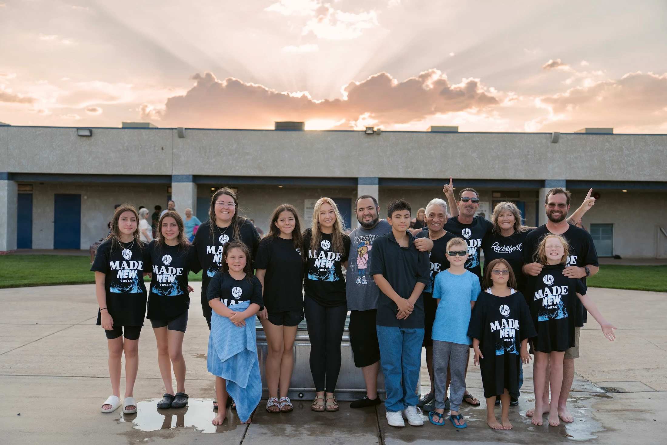 A large, multi-generational group smiles for a photo outdoors, many wearing matching 'Made New' t-shirts after a baptism, with a sunset in the background.
