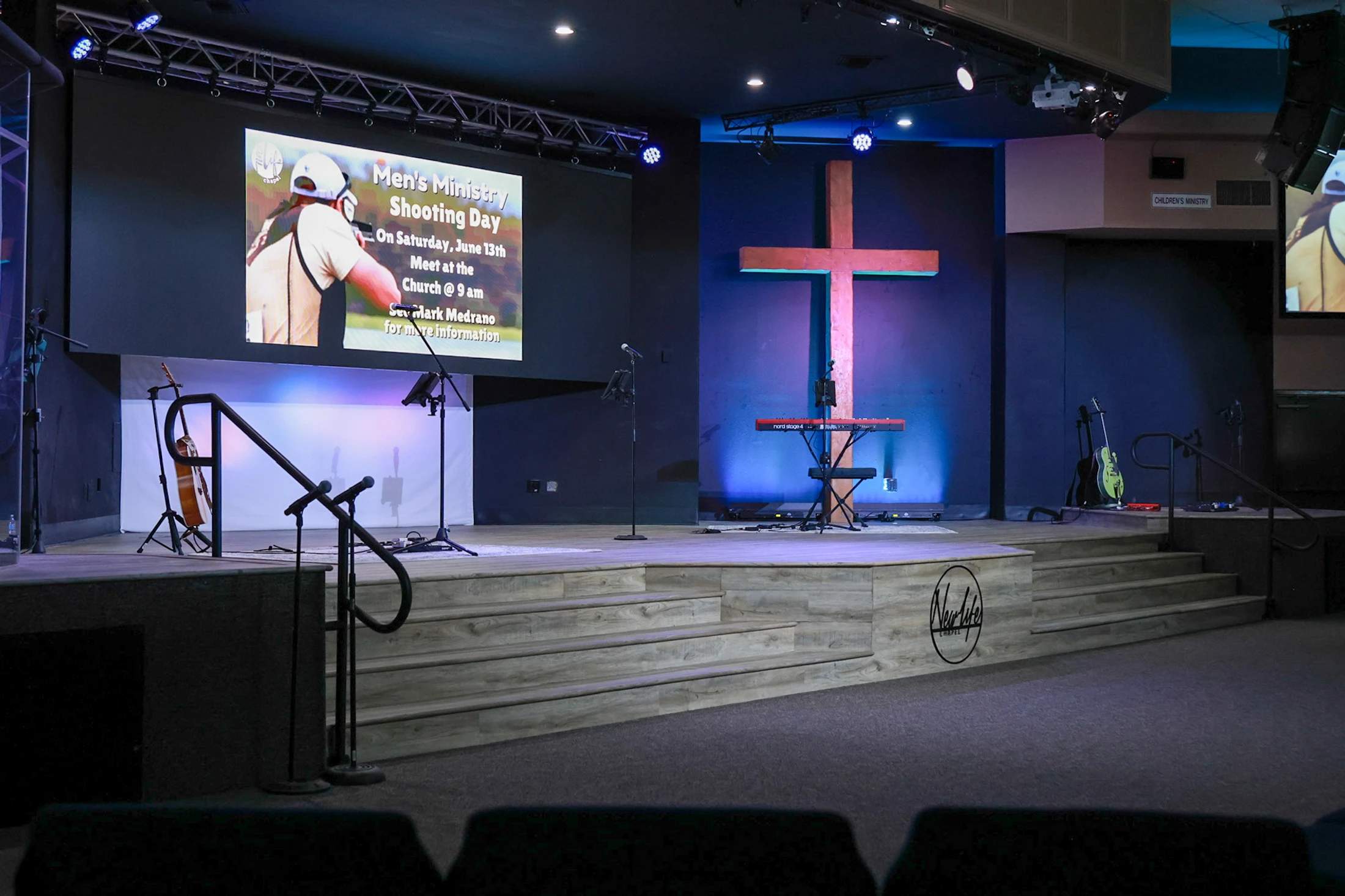 An empty stage in a modern church features a large wooden cross, musical instruments, and a screen advertising a "Men's Ministry Shooting Day."