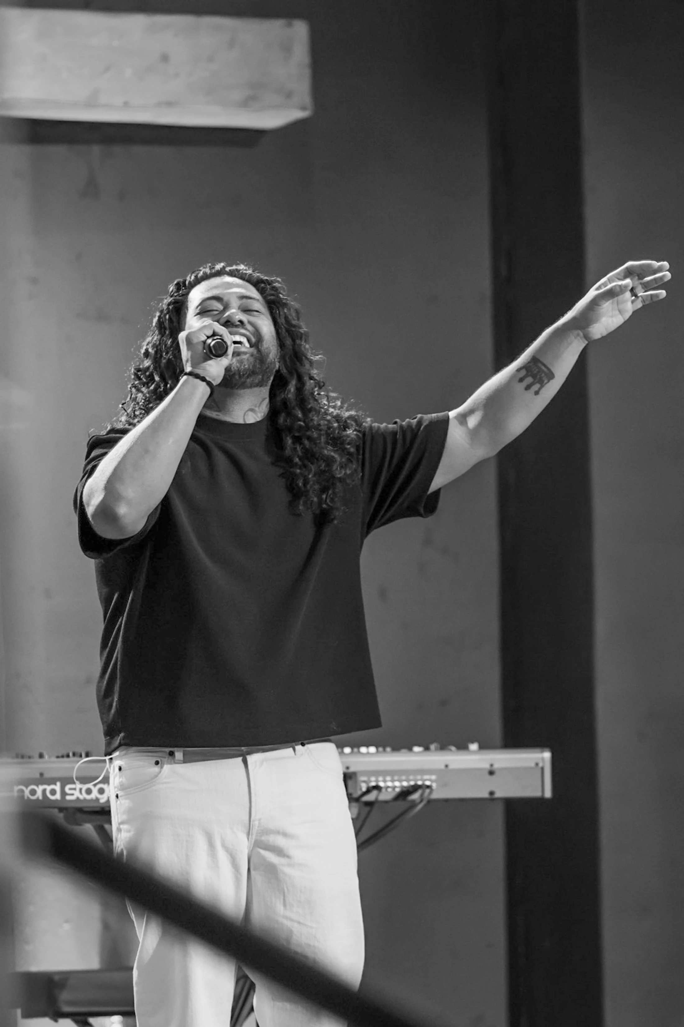 A black and white photo of a man with long curly hair and a beard singing into a microphone, with his eyes closed and one arm raised in the air.