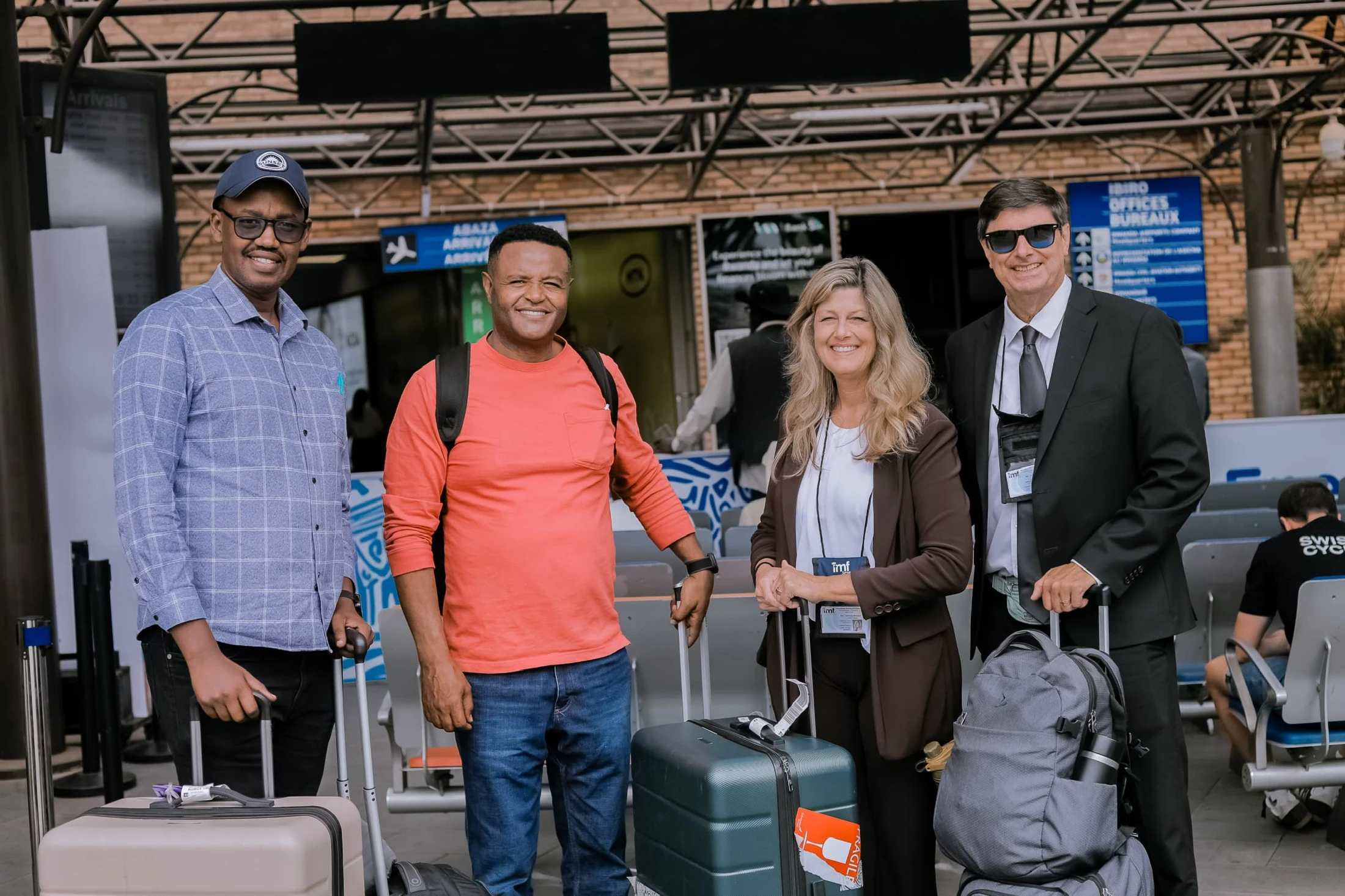Four diverse travelers, three men and one woman, smile for a photo with their luggage after arriving at an airport terminal.