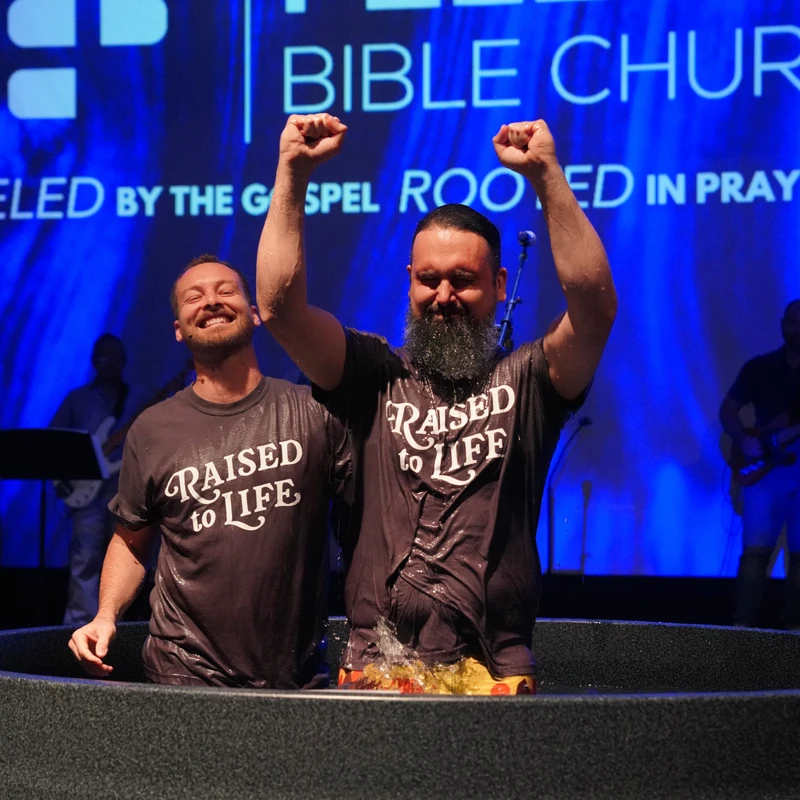 A bearded man joyfully raises his arms after being baptized in a church pool. Another man smiles beside him. Both wear 'Raised to Life' T-shirts.