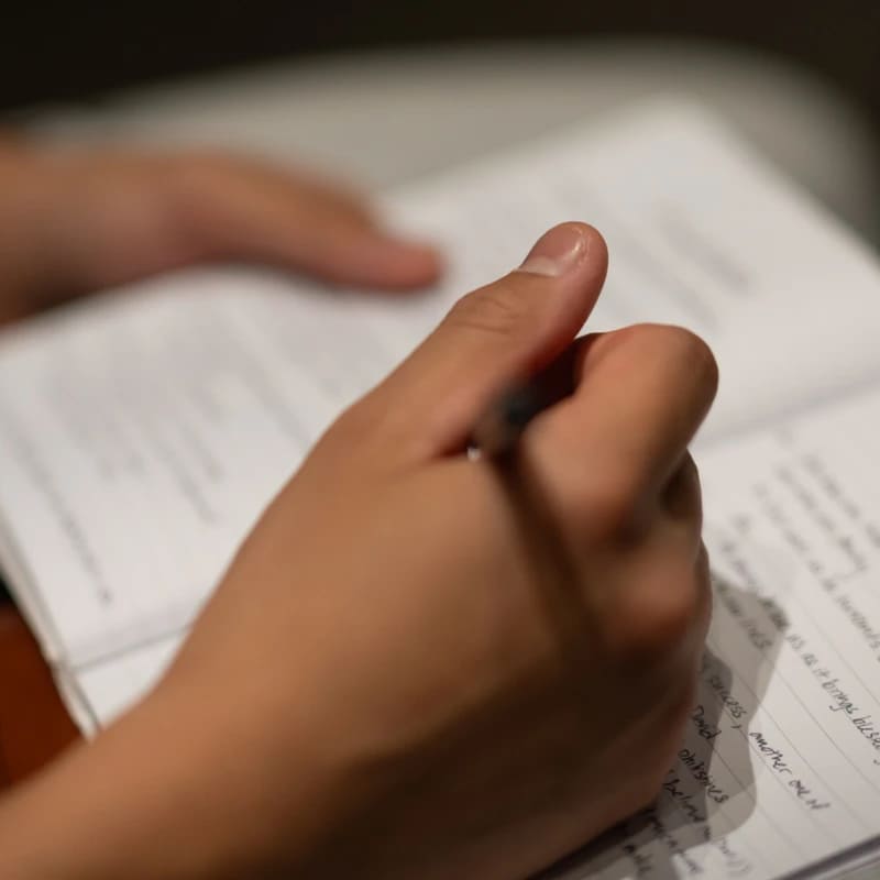 A person's hand holds a pen and writes in a lined notebook. The focus is on the hand, with the handwritten text and background blurred.