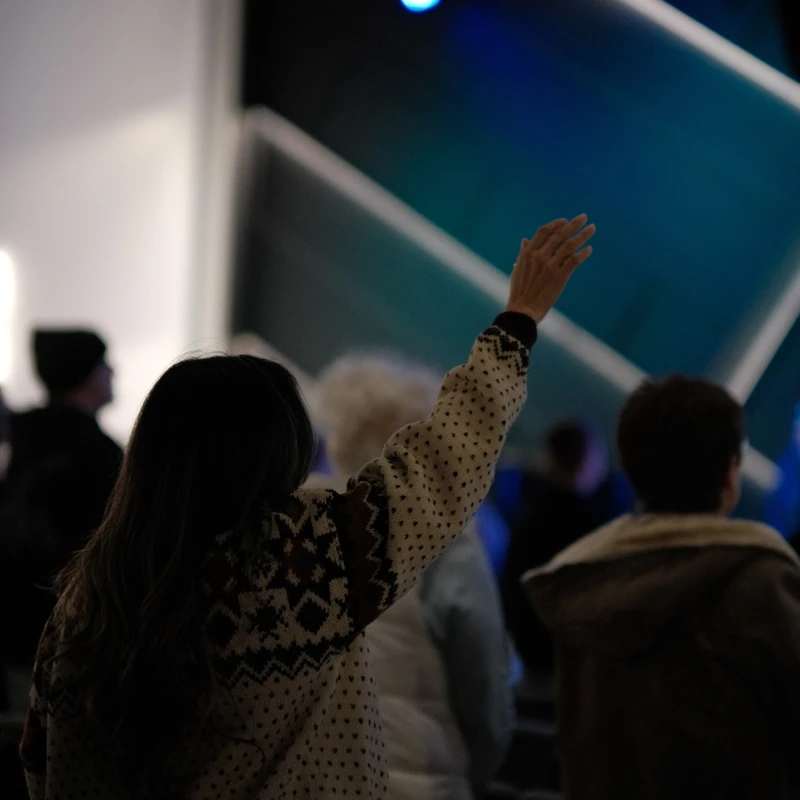 A woman seen from behind in a patterned sweater raises her left hand during a service or concert, with a softly lit blue stage in the background.