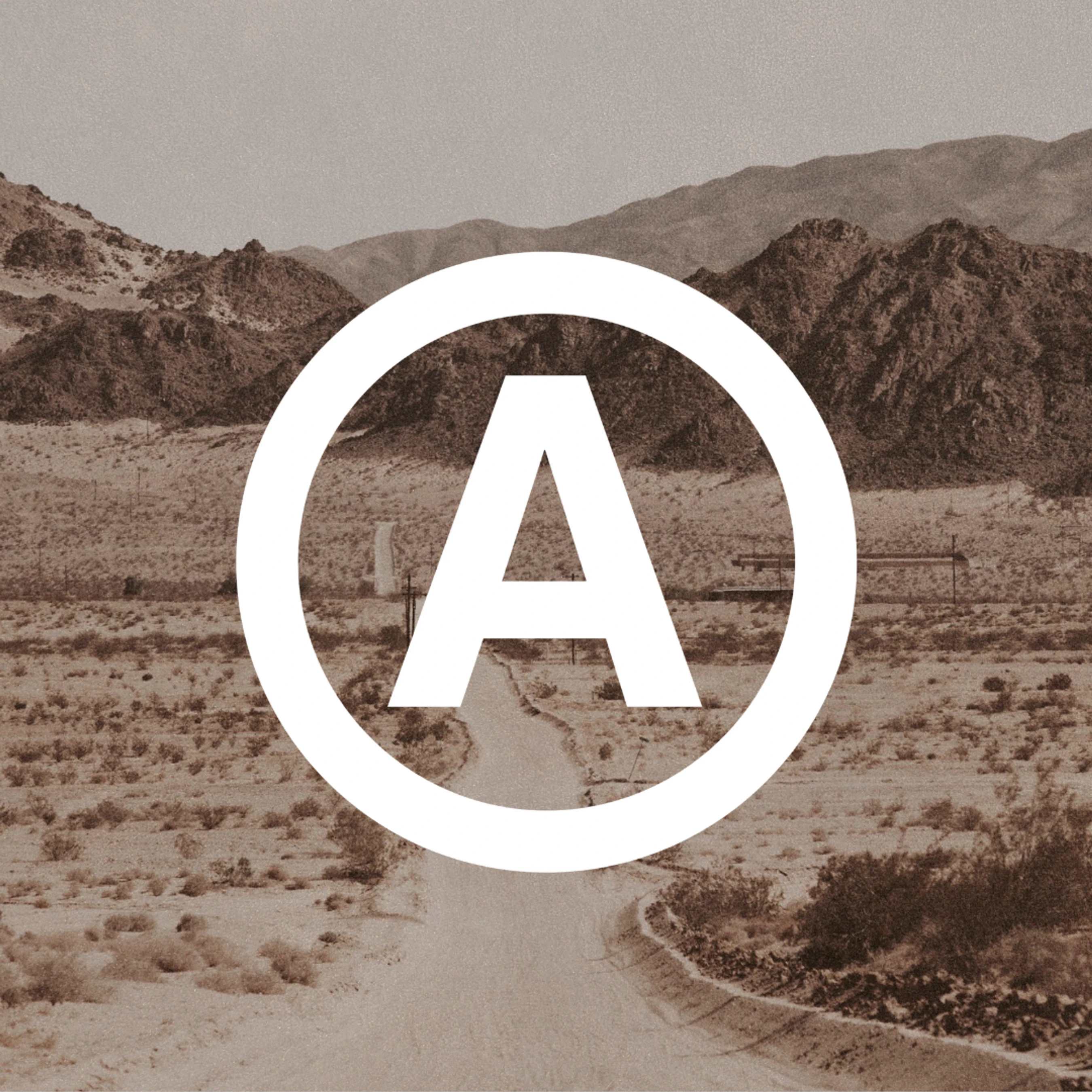 A white capital letter 'A' inside a circle is centered over a sepia-toned photo of a winding dirt road in a desert with mountains in the background.