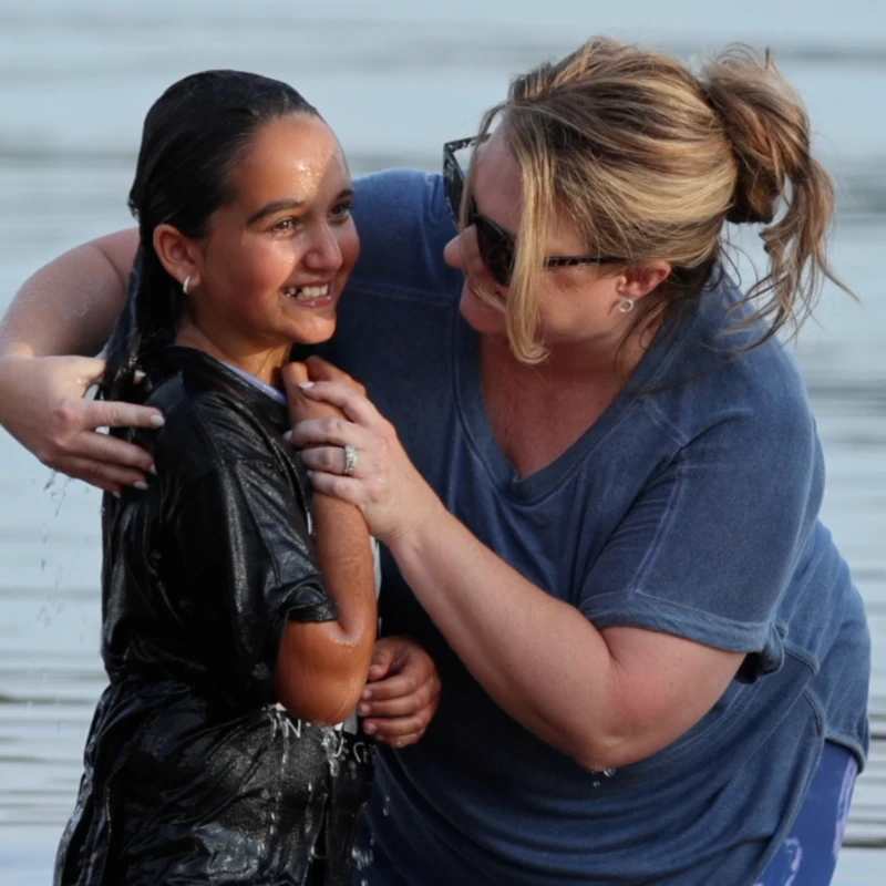 A woman in a blue shirt smiles as she holds a young girl in a wet black shirt in the water during a baptism. The girl smiles back at her.