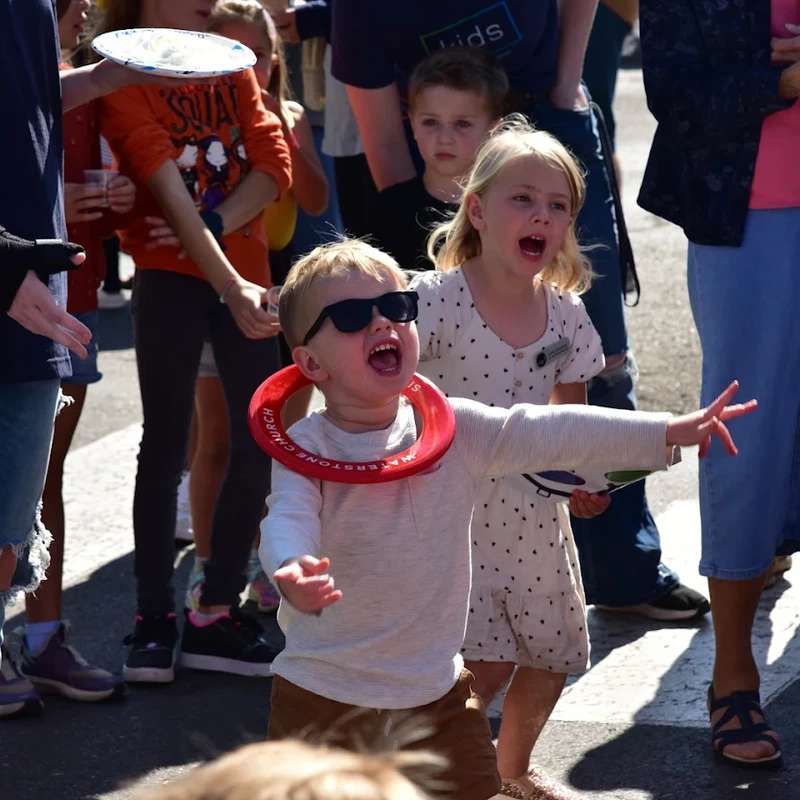 A young boy in sunglasses with a red ring around his neck shouts with joy, surrounded by other children and adults at a sunny outdoor gathering.