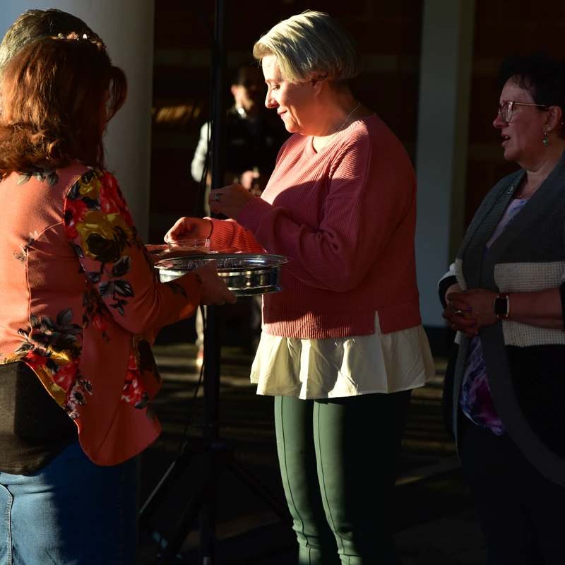 A woman in a pink sweater receives communion from a tray held by another woman. A third woman stands nearby, all bathed in a warm ray of sunlight.