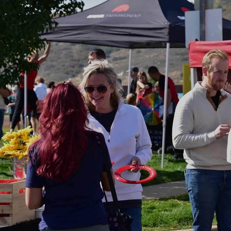 A blonde woman holding a red frisbee smiles while talking to a woman with red hair at a sunny outdoor community event with tents in the background.