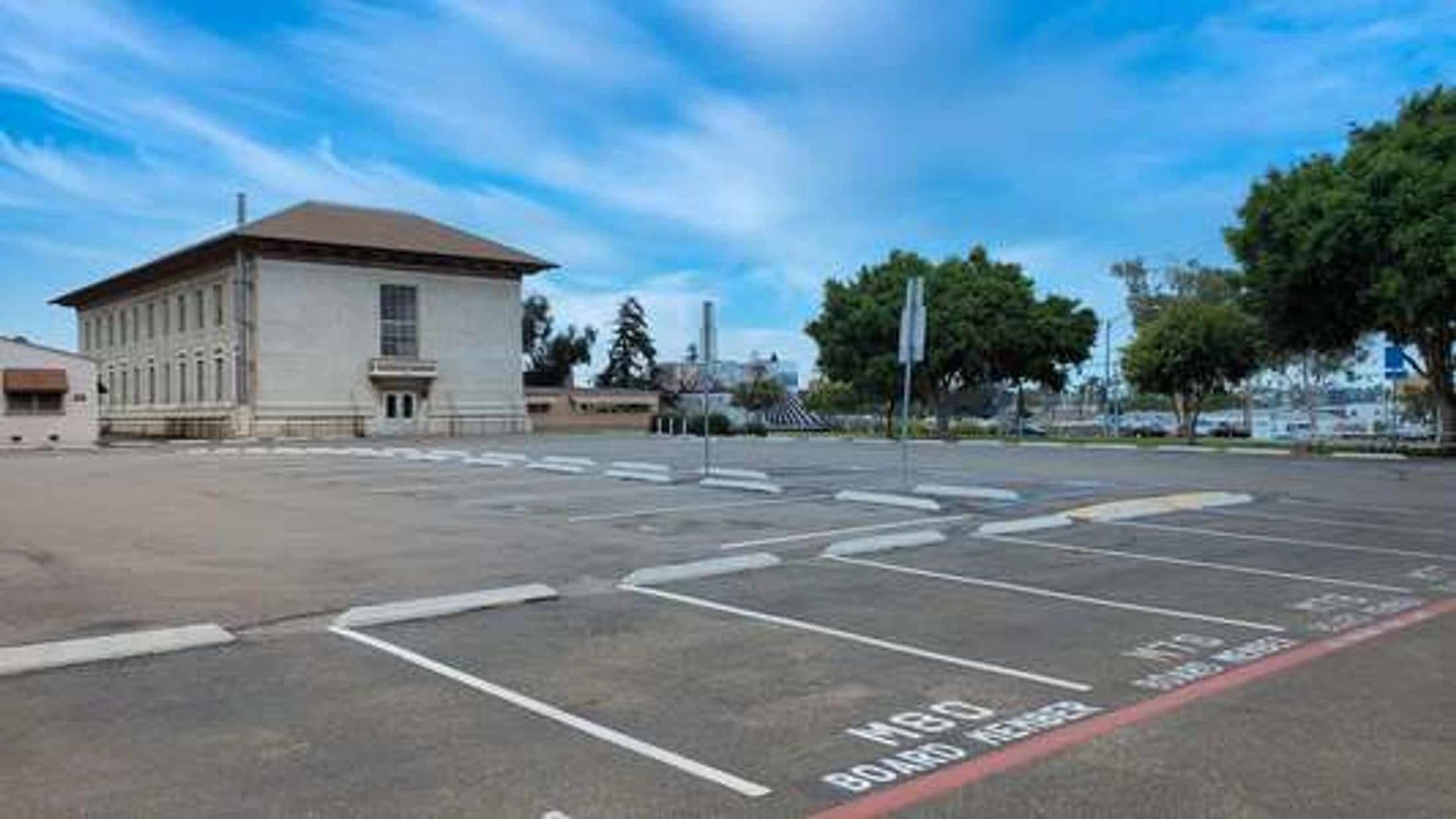 An empty asphalt parking lot sits in front of a large, two-story building under a blue sky. A foreground parking space is marked for a "Board Member."