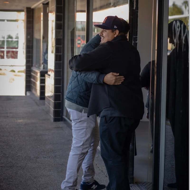 Two men share a friendly hug on a sidewalk in front of a building with large glass windows. The man on the right wears a baseball cap and smiles warmly.
