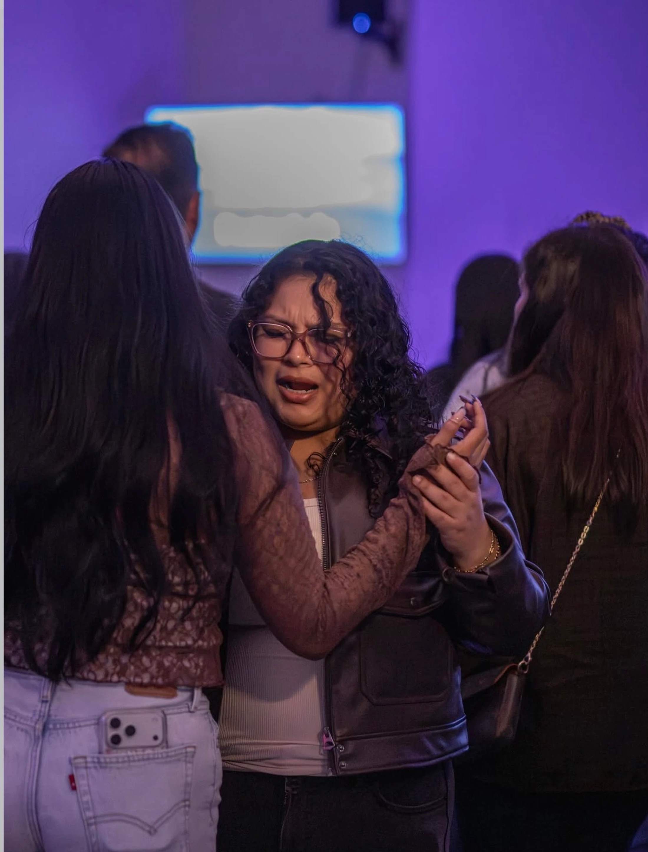 A young woman with glasses and curly hair sings with intense emotion while another woman, seen from behind, holds her hand in a room with purple lighting.