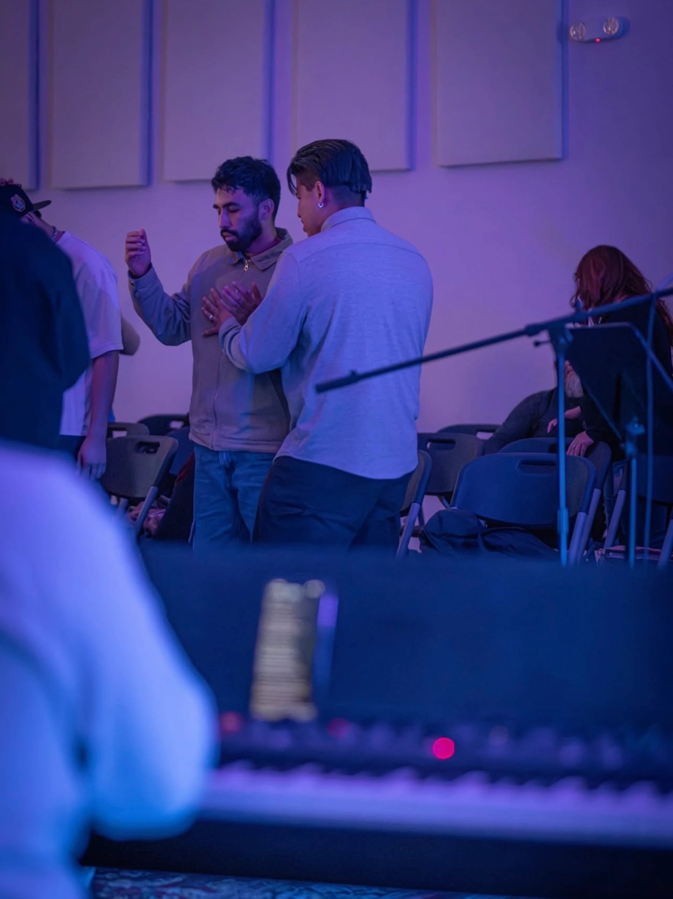 Two men stand together in prayer in a room with blue lighting. One man has his hand on the other's chest in a supportive gesture during a worship gathering.